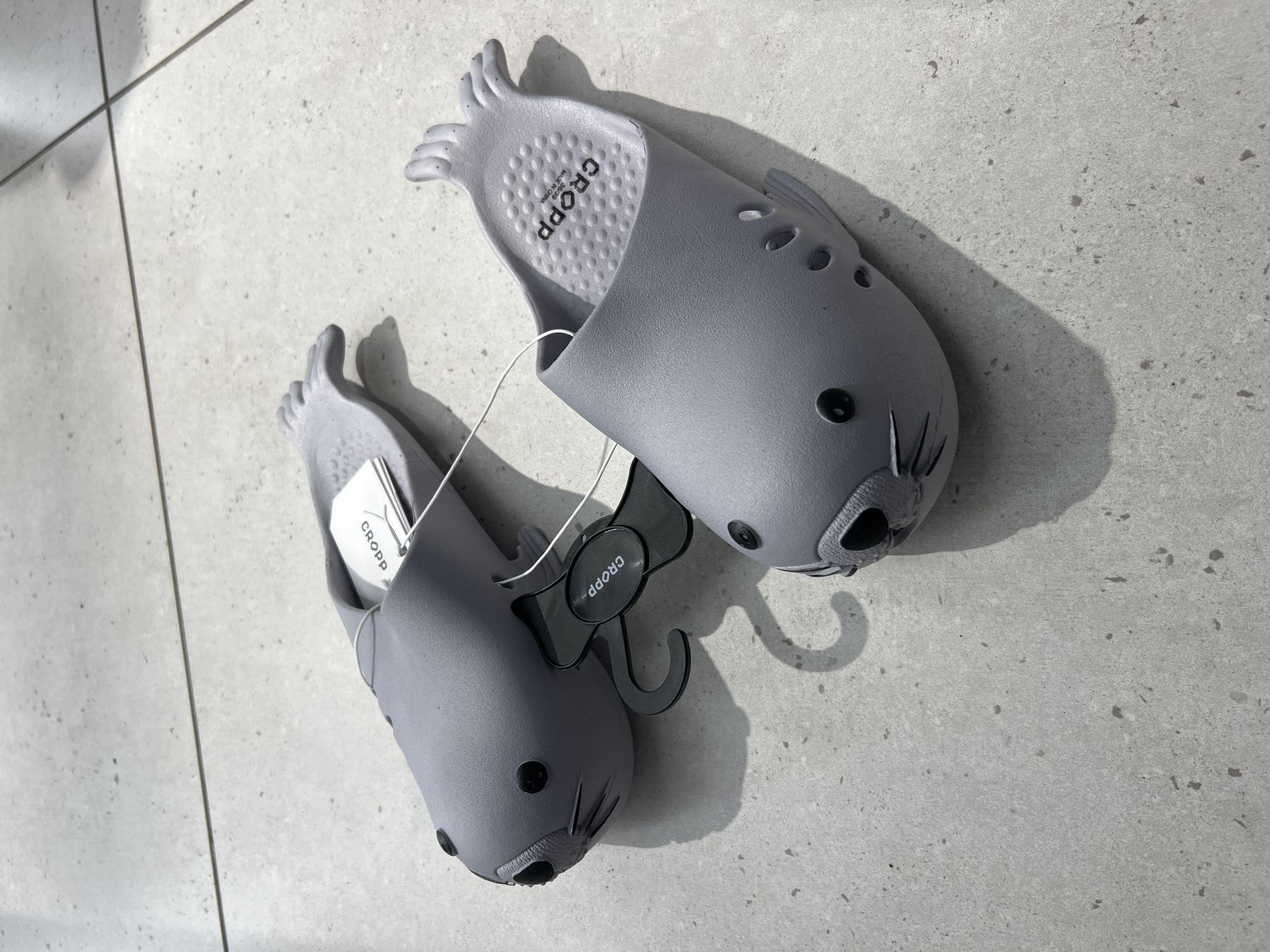 Two playful, animal-themed slippers are depicted from an overhead angle on a light grey, speckled tiled floor. The slippers, likely made of EVA foam or a similar soft material, are a muted grey color and designed to resemble seals or other marine creatures, featuring distinct eyes, snouts, and etched whiskers. The heel area of the slippers has a subtle, textured, scale-like pattern, while the front part is shaped like small flippers or toes.
One slipper is positioned with its sole facing upwards, revealing branding and manufacturing information. The brand name "CROPP" is prominently visible in capital letters on the insole, along with the text "MADE IN CHINA" and the size "38/39". Both slippers are connected by a thin white string and feature a black plastic hanger, also bearing the "CROPP" logo. Small white tags with the "CROPP" logo are attached to the slippers, suggesting they are new and likely displayed for sale in a retail environment. The floor is composed of large, light-colored tiles with darker speckles and clear grout lines, suggesting an indoor setting. The lighting is bright and even, casting subtle shadows from the slippers. This image was taken in Belgrade, Serbia.