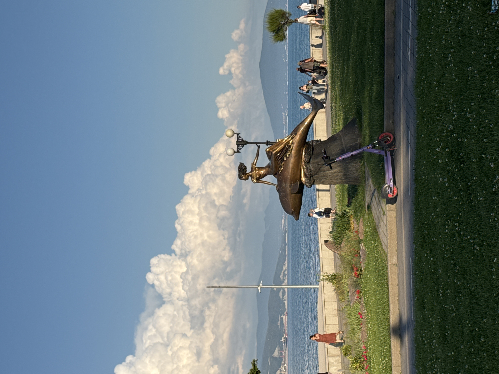 A bronze sculpture of a woman with flowing hair sitting on a dolphin is prominently displayed in a park setting. The sculpture is positioned on a stone base with a lamppost behind it. To the left of the sculpture, a woman in a reddish-brown dress walks along a paved pathway. Further down the pathway, a group of people, including children, are visible, some walking and others standing. A purple electric scooter is parked on the grass to the left of the pathway. In the background, a body of water, likely the sea, stretches towards distant mountains. The sky above is a clear blue with large, fluffy white clouds. The park area features green grass, some scattered flowers, and a few trees. The overall scene depicts a public space by the water, with people enjoying a leisurely day outdoors.