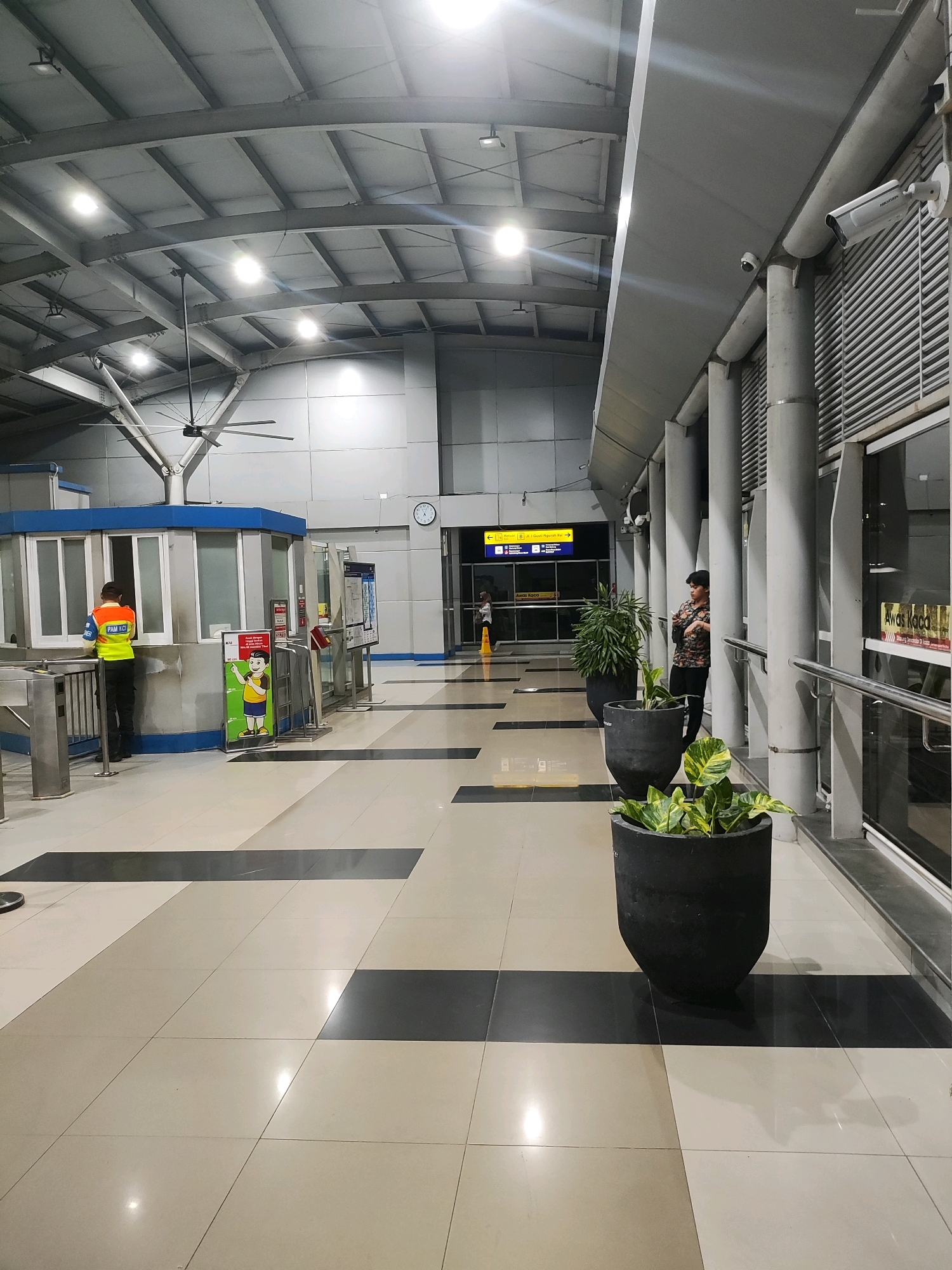 The image depicts the interior of a modern, well-lit train station in East Jakarta, Indonesia. The spacious concourse features a high, vaulted ceiling with exposed metal beams and numerous fluorescent light fixtures. The floor is tiled in a pattern of beige and dark gray rectangles.
On the left side of the image, a security guard in a bright orange and yellow vest stands behind a blue and gray ticket booth with large glass windows. A metal turnstile is visible in front of the booth. A colorful promotional poster featuring a cartoon character is displayed next to the booth.
Further down the concourse, large cylindrical concrete columns support the elevated ceiling. Two large black planters containing lush green plants are positioned along the right side of the walkway. A person in a patterned shirt is standing near the planters, looking towards the right.
In the background, a sign with yellow text and arrows indicates directions to different platforms. A clock is mounted on the wall behind the security booth. Security cameras are visible mounted on the ceiling and on the upper portion of the wall. The overall impression is one of a clean, organized, and functional public transportation hub.