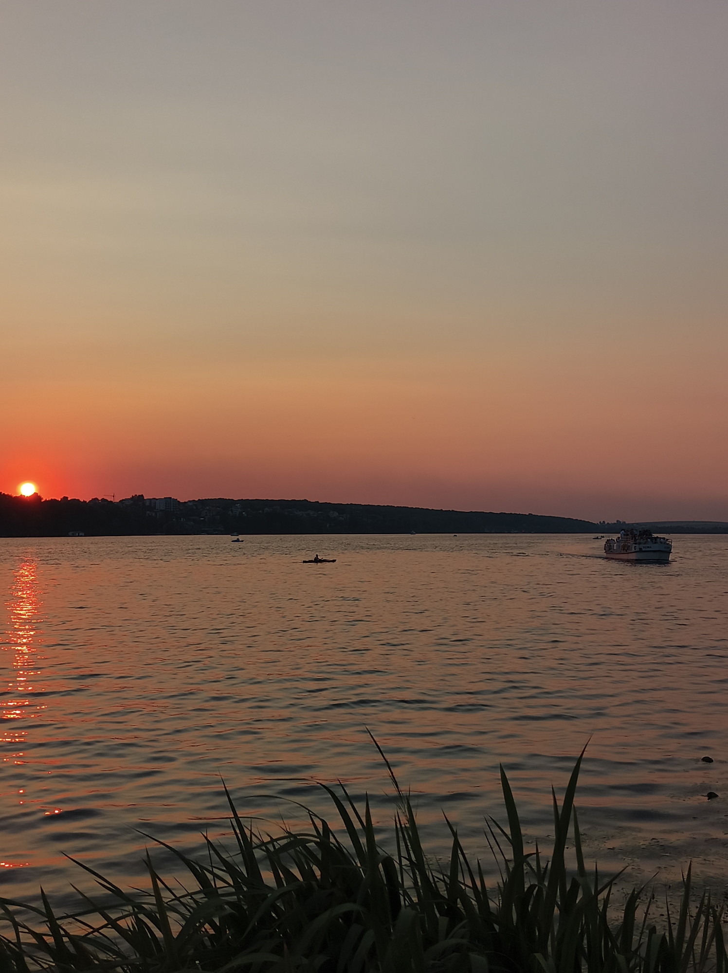 The image depicts a sunset over Ternopil Lake in Ternopil, Ukraine. The sun, a bright orange orb, is setting on the left side of the frame, casting a warm glow across the sky and reflecting on the water's surface. The sky transitions from a pale orange near the horizon to a muted grey higher up.
A silhouetted treeline with buildings and a crane marks the distant shore of the lake. Several small boats are visible on the water. In the mid-ground, a single person is kayaking. Further to the right, a larger boat, likely a tour boat, is carrying passengers. The water itself is rippled, with the sunlight creating shimmering patterns. In the foreground, dark green reeds or tall grass are visible, adding depth to the scene. The overall atmosphere is serene and tranquil, characteristic of an evening by the lake.