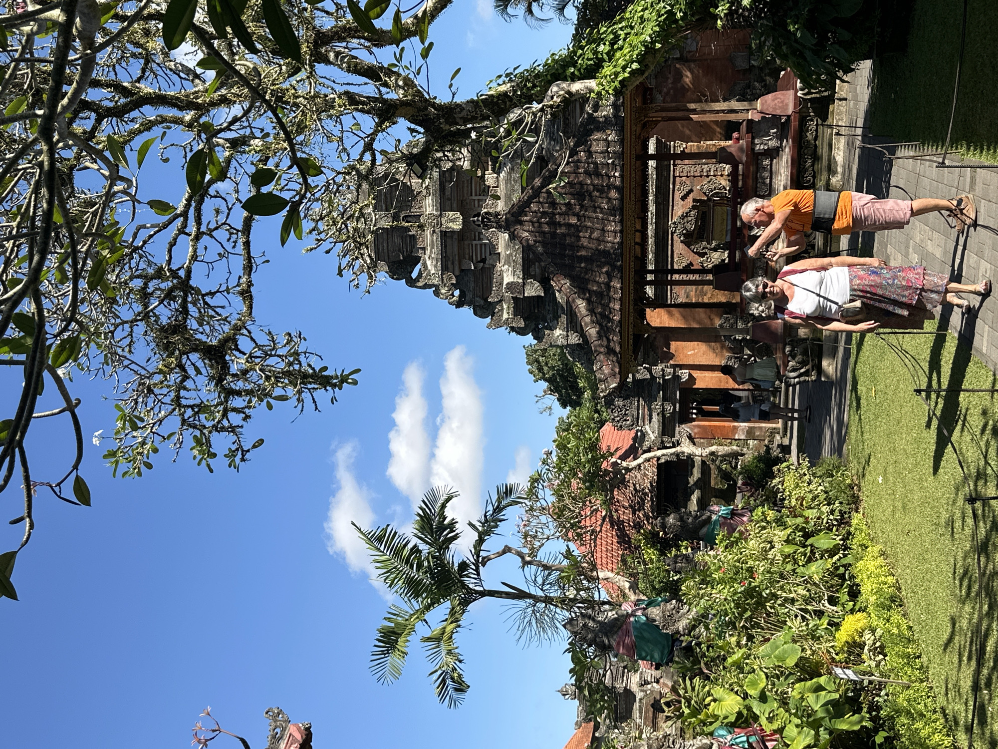 In Ubud District, Indonesia, two tourists walk on a paved path in front of a Balinese temple. The woman in the foreground wears a white tank top, a patterned skirt, sunglasses, and a backpack, while the man beside her is dressed in an orange t-shirt, pink shorts, and a grey back support. He holds up a phone, possibly taking a picture. The temple features traditional tiered roofs, intricate carvings, and is surrounded by lush greenery, including palm trees and flowering bushes. A large, ancient tree with moss-covered branches looms over the scene, framing the blue sky with scattered clouds. The sunlight casts shadows on the well-maintained lawn, indicating a bright, sunny day. Several other people can be seen in the background, suggesting the temple is a popular tourist destination.
