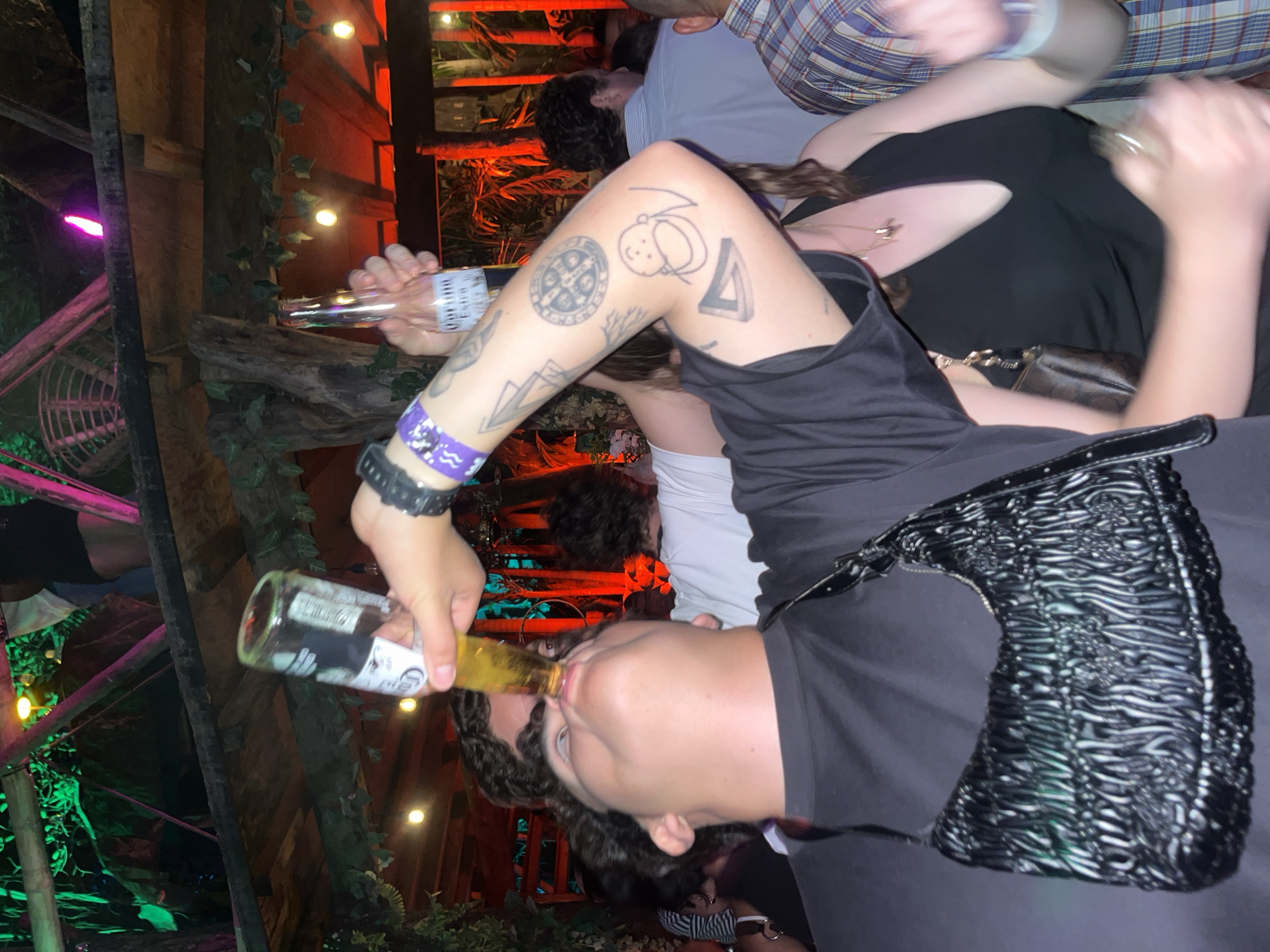 The image depicts a person drinking from a Corona bottle. The person is wearing a black shirt and has tattoos on their left arm. They also have a black watch on their left wrist and a purple wristband. A black, patterned bag is visible on their shoulder. Other people are in the background, including one wearing a plaid shirt. The location appears to be outdoors, possibly at a bar or event space, with foliage and string lights visible in the background. There are wooden structures and decorative elements such as a pink fan in the background. Given the prompt’s instruction to specify the location as Brisas de Zicatela, Mexico, the image is set within that context.