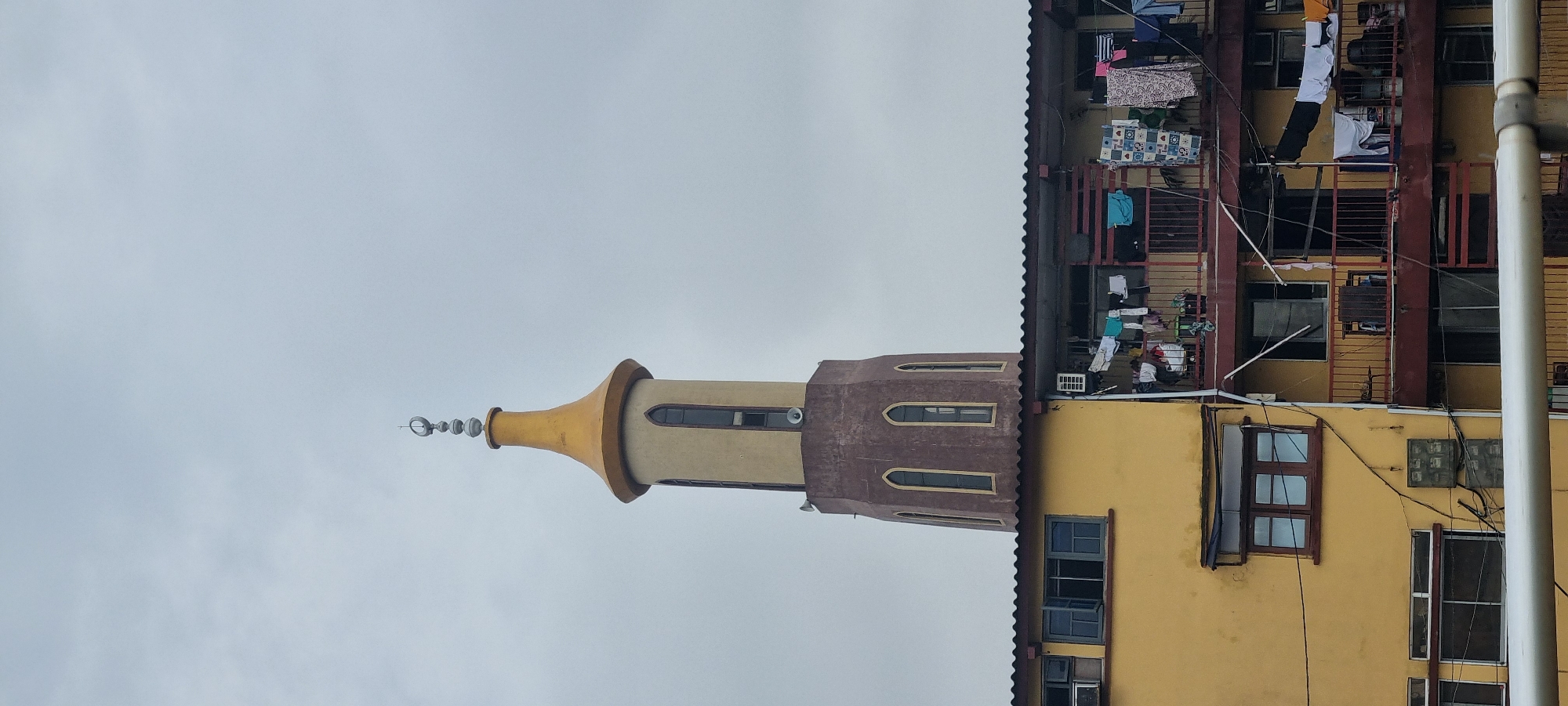 The image shows a minaret of a mosque next to a multi-story residential building in the city of Unknown, Unknown. The minaret is two-toned, with a gold, bell-shaped top section and a brown base section with arched windows. It is topped with a crescent moon finial. The building has a yellow exterior wall with brown window frames. The upper floors of the building have balconies with clothes hanging on them. The sky is overcast.