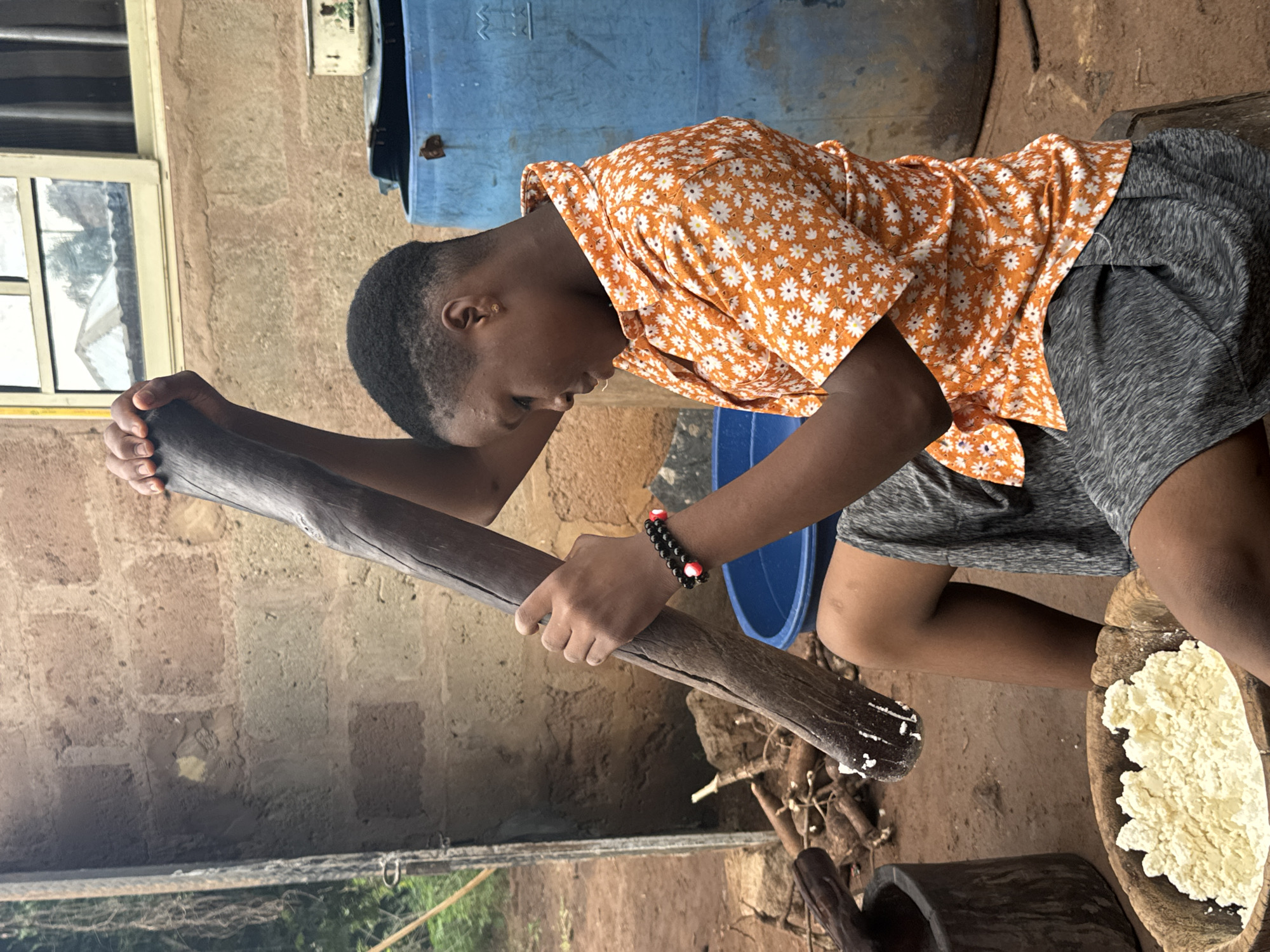 In Eziudo, Nigeria, a young person is seated outdoors, engaged in the task of pounding yam. They are positioned near a wooden mortar filled with cooked yam. The person is wielding a large wooden pestle, using it to mash the yam in the mortar. They are wearing an orange floral shirt and grey shorts. A blue plastic barrel is visible in the background. The surrounding environment appears to be a rural or semi-rural setting, possibly a courtyard or backyard. The construction of the building in the background is a rough brick structure with a visible window. There are also some dry branches and wooden pieces on the ground.