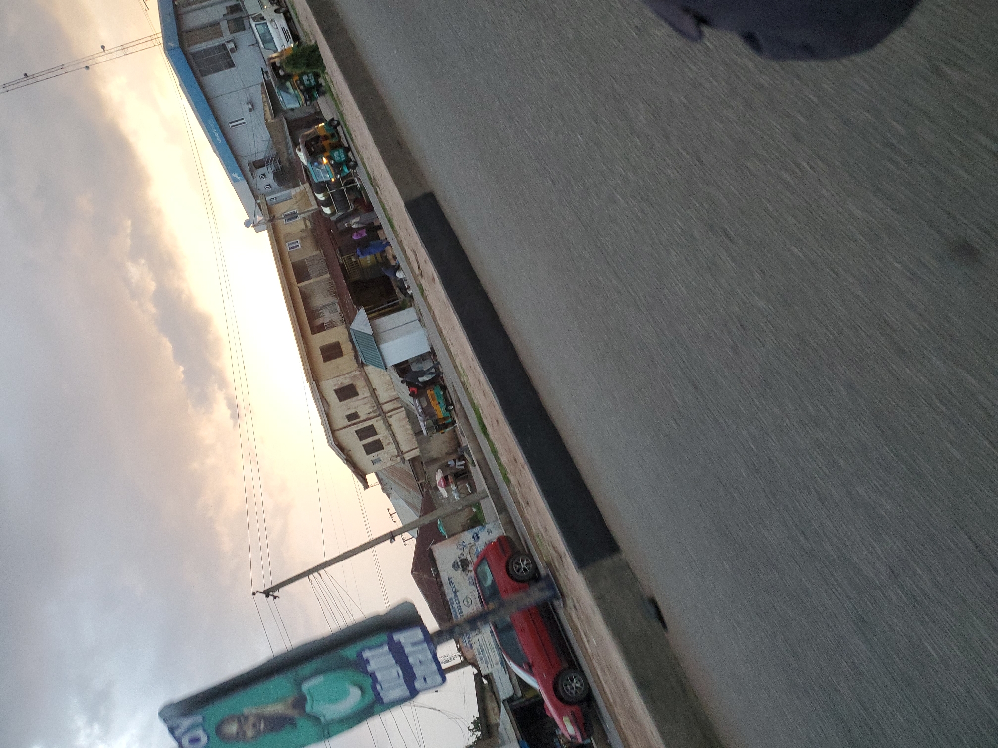 The image depicts a street scene in Jos, Nigeria, likely during the late afternoon or early evening, given the soft, diffused light and the golden hues in the sky. The perspective is from a moving vehicle, creating a slight blur and tilt.
In the foreground, a grey asphalt road with a dark grey concrete curb is visible, indicating a built-up area. To the left, a tall, green and blue billboard stands tilted, featuring a man's face and text that includes "Right Guard."
Further down the street, on the right side of the road, a line of buildings is situated. The building closest to the center is a light-colored structure with multiple windows and balconies. To its right, a modern building with a blue facade and "Union Bank" signage stands prominently, topped with a tall communication tower.
Several vehicles are present. A red station wagon is parked on the side of the road in front of the buildings. Further along, three auto-rickshaws, typical modes of transport in Nigeria, are parked or driving slowly. One is green and yellow, another is primarily yellow, and a third is green. A white utility vehicle is parked near the Union Bank building.
A few people are visible on the sidewalks and in front of the shops. They appear to be engaged in various activities, such as standing, talking, or tending to businesses. The general atmosphere suggests a bustling commercial area. The sky is partly cloudy, with soft orange and grey tones indicating sunset or sunrise. The overall scene captures a slice of everyday life on a busy street in Jos.