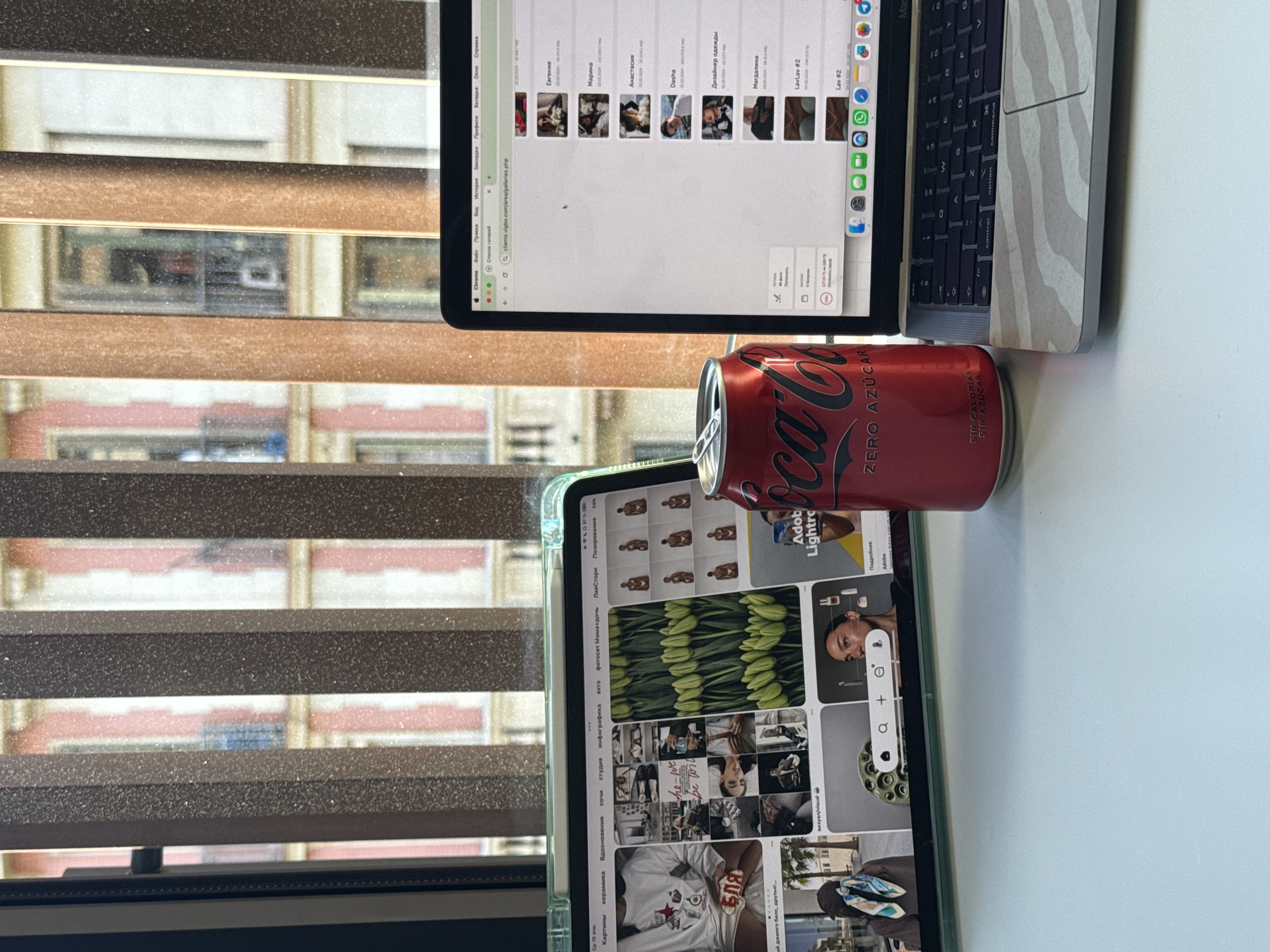 The image shows a desk setup with a laptop, a tablet, and a can of Coca-Cola Zero. The laptop is open, displaying a Chrome browser window with multiple images, likely related to a photo gallery or design project. The tablet also has an image-heavy page open, with various photos and designs, potentially from a visual inspiration platform. The Coca-Cola Zero can is red with Spanish text. The background features a window with blinds, and outside the window, there's a blurred view of buildings, with beige/brown/red brick facade, typical of Barcelona.