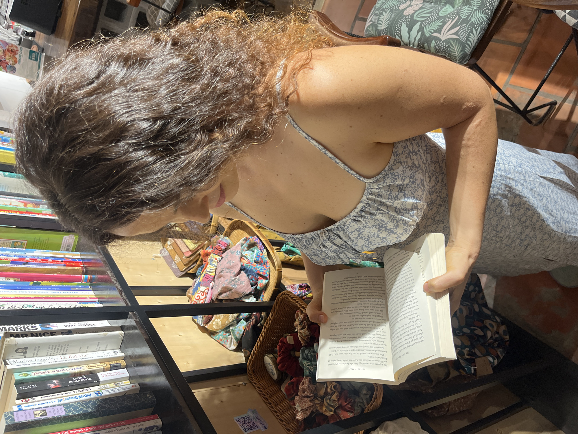 A woman with curly brown hair is reading a book. She is wearing a light blue and white floral print dress. The book is open and she is holding it with both hands. In the background, there is a shelf filled with books. To the right of the bookshelf, there is a wicker basket filled with colorful fabric scraps. The scene appears to be indoors, possibly in a shop or a home. The lighting suggests it might be daytime, with sunlight streaming in from the side. The image does not contain any elements that can identify the location as Phu Thuy Ward, Vietnam.