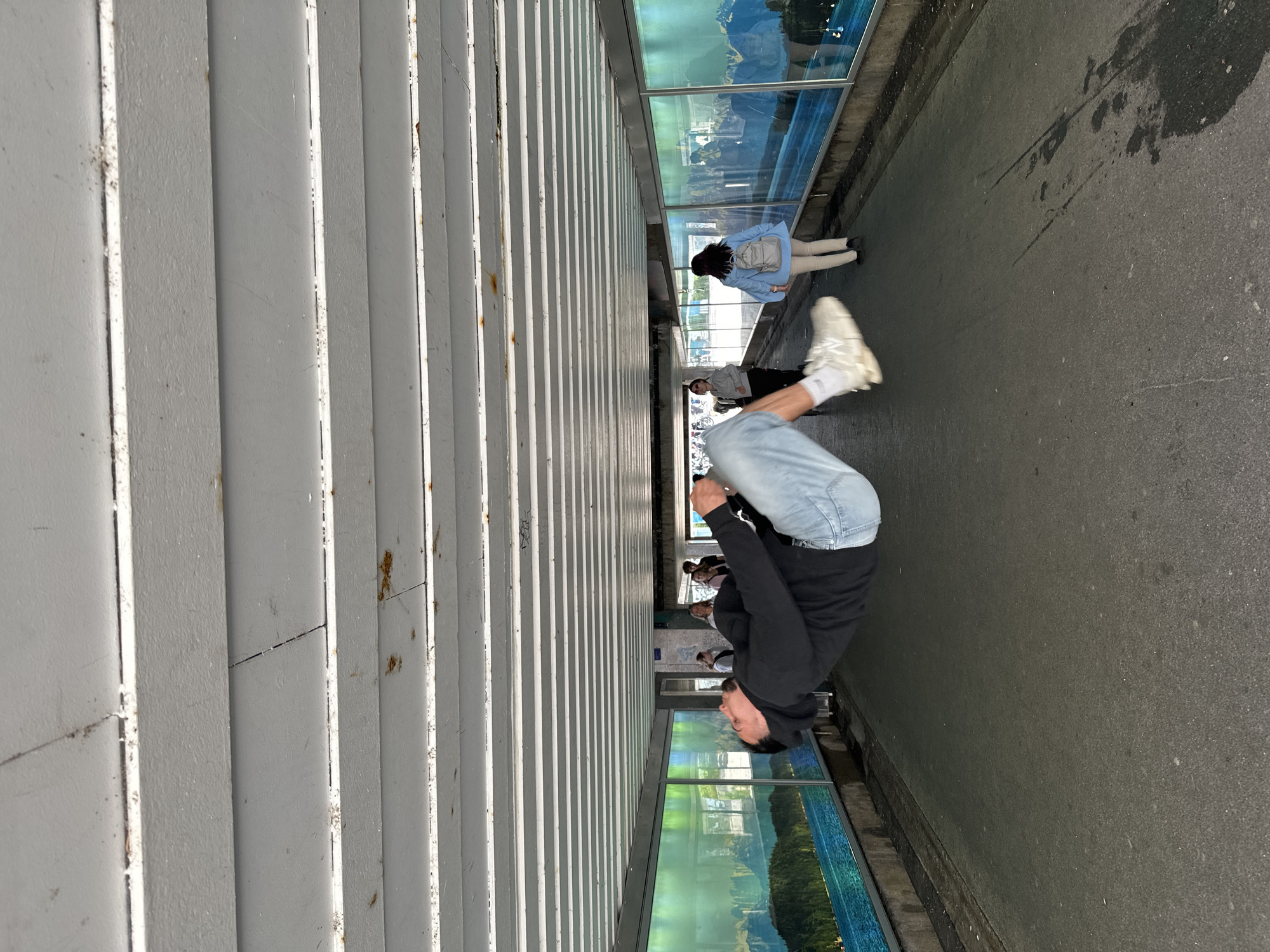 A young man is captured mid-air, appearing to float horizontally across a pedestrian walkway under a covered structure. He is wearing a black hoodie, blue jeans, and white sneakers. His body is bent at the waist, with his legs tucked towards his chest. In the background, several other people are walking in both directions on the walkway. The walkway is lined with large, panoramic murals depicting a scenic landscape of water, trees, and mountains, possibly a reflection of the natural beauty surrounding Starnberg, Germany. The ceiling of the walkway is constructed with horizontal white beams. The overall scene suggests an urban environment, possibly a pedestrian underpass or a covered street, enhanced by artistic murals.