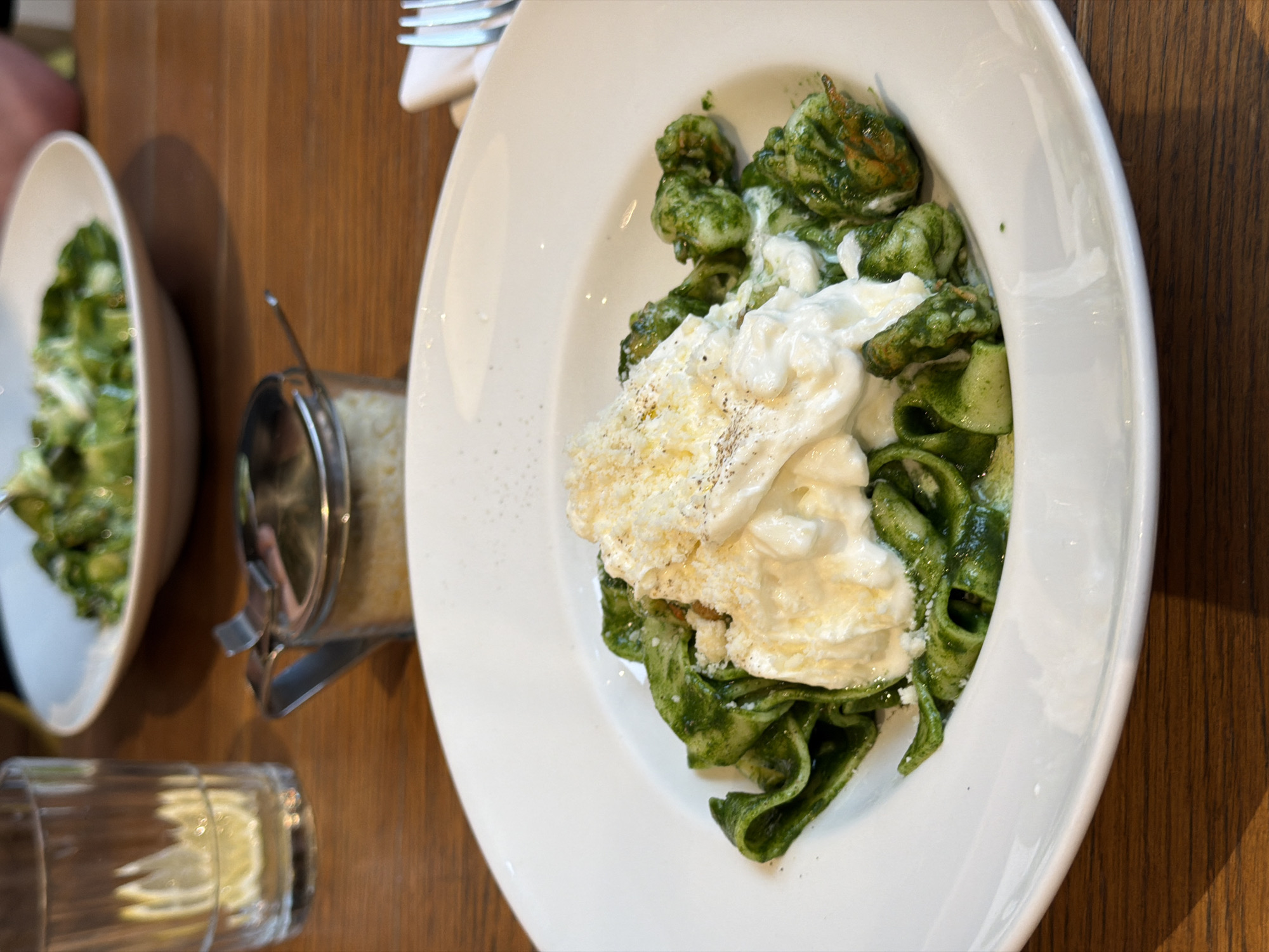 A high-angle shot shows a plate of green pasta topped with white cheese and grated parmesan, a small bowl of green food, a parmesan cheese shaker, a glass of water with a lemon slice, and silverware on a wooden table. The environment suggests a restaurant setting. The city of Moscow, Russia, cannot be definitively determined from the image.