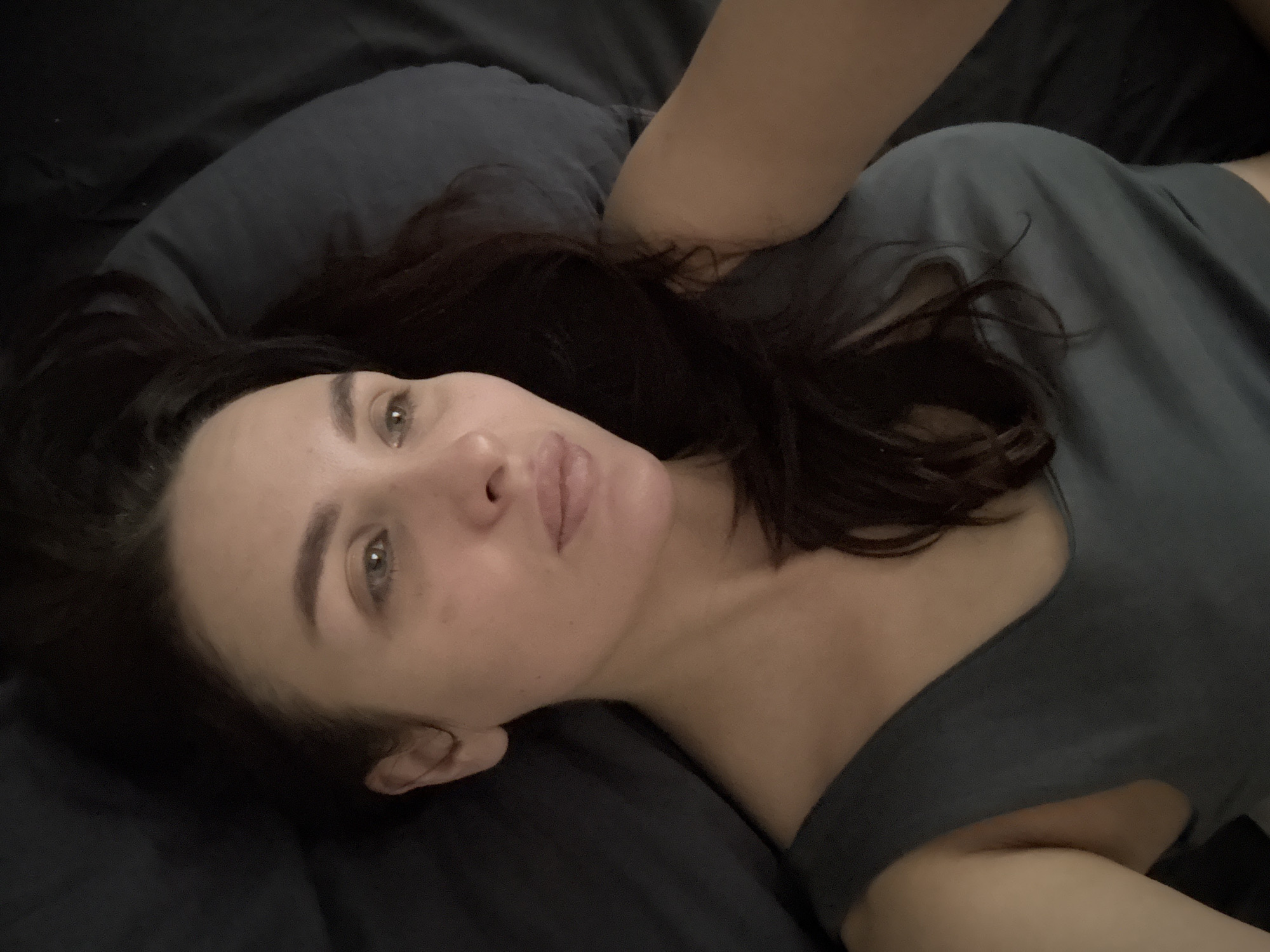 A woman with dark hair and light eyes is lying down, possibly on a bed, given the dark-colored pillow and blanket/sheet beneath her. She is wearing a gray top. The setting appears to be indoors, given the lighting and domestic elements present. No information indicates that the photo was taken in Chaikovsky, Russia.