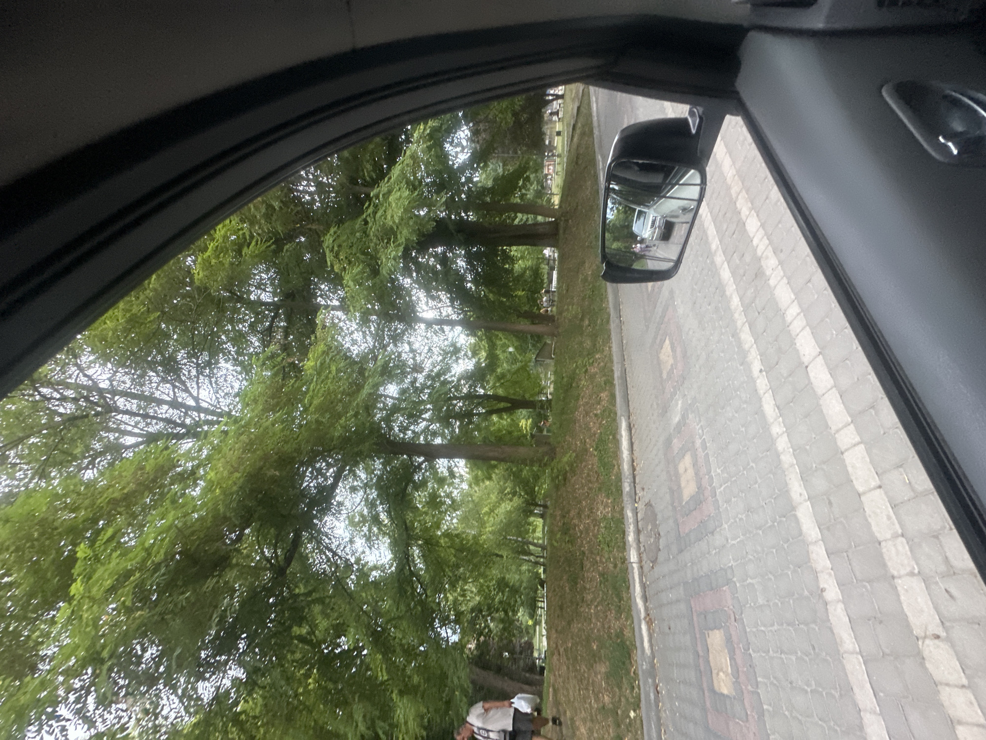 The image captures a view from inside a vehicle looking outwards through the side window, with a side mirror reflecting a street scene. The scene depicts a tree-lined park area with a paved walkway or road. In the foreground, the textured pavement of the walkway features a pattern of square inlays of different colors. The side mirror shows a silver car and two pedestrians walking on the paved area. Beyond the walkway, a grassy embankment rises to a park filled with lush green trees. Several people are visible sitting on benches or walking in the distance within the park. The overall environment suggests a public park or recreational area within a city, likely Kamianets-Podilskyi, Ukraine, based on the context provided. The lighting indicates it is daytime, with overcast conditions.