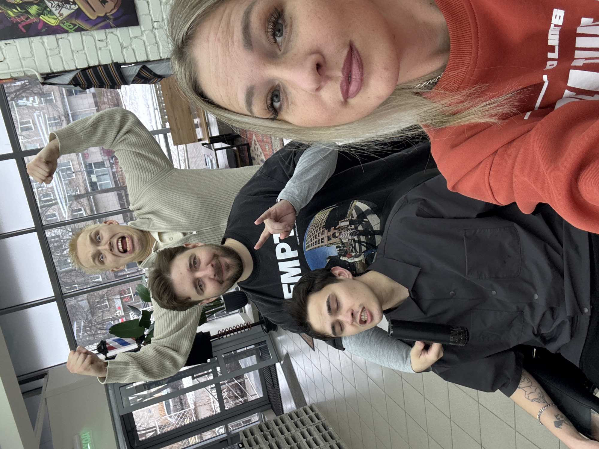 Four individuals are captured in a selfie taken in Chaikovsky, Russia. The foreground shows a blonde woman wearing a red sweatshirt. Behind her, a man in a black t-shirt makes a hand gesture. Further back, a person with blonde hair is seen near a window, posing with arms raised. Another individual is positioned beside the man in the black t-shirt. The scene is inside a building with large windows showing a cityscape, and the interior features tiled floors and seating.