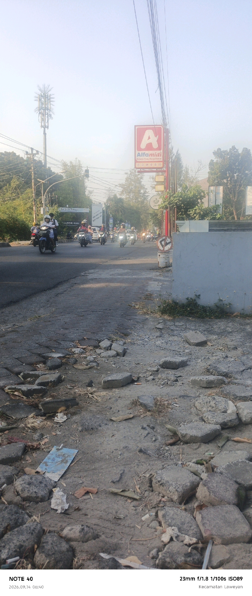 This image depicts a street scene in the Laweyan subdistrict of Surakarta, Indonesia, at approximately 6:40 AM on September 14, 2026. The foreground is dominated by a broken and uneven asphalt road surface littered with debris, including broken concrete, stones, dried leaves, and discarded paper and plastic. In the midground, a group of motorcycles are riding down the road, many with riders wearing helmets. Some riders appear to be students, indicated by their uniforms. To the right of the road, a white concrete wall runs along the edge of the frame. Further back, a large red and white sign for "Alfamidi" is visible, a common convenience store chain in Indonesia, along with other businesses and trees lining the street. A cell tower stands tall against the hazy sky. The overall impression is of a busy urban street at the beginning of the day, with visible signs of disrepair on the road surface.