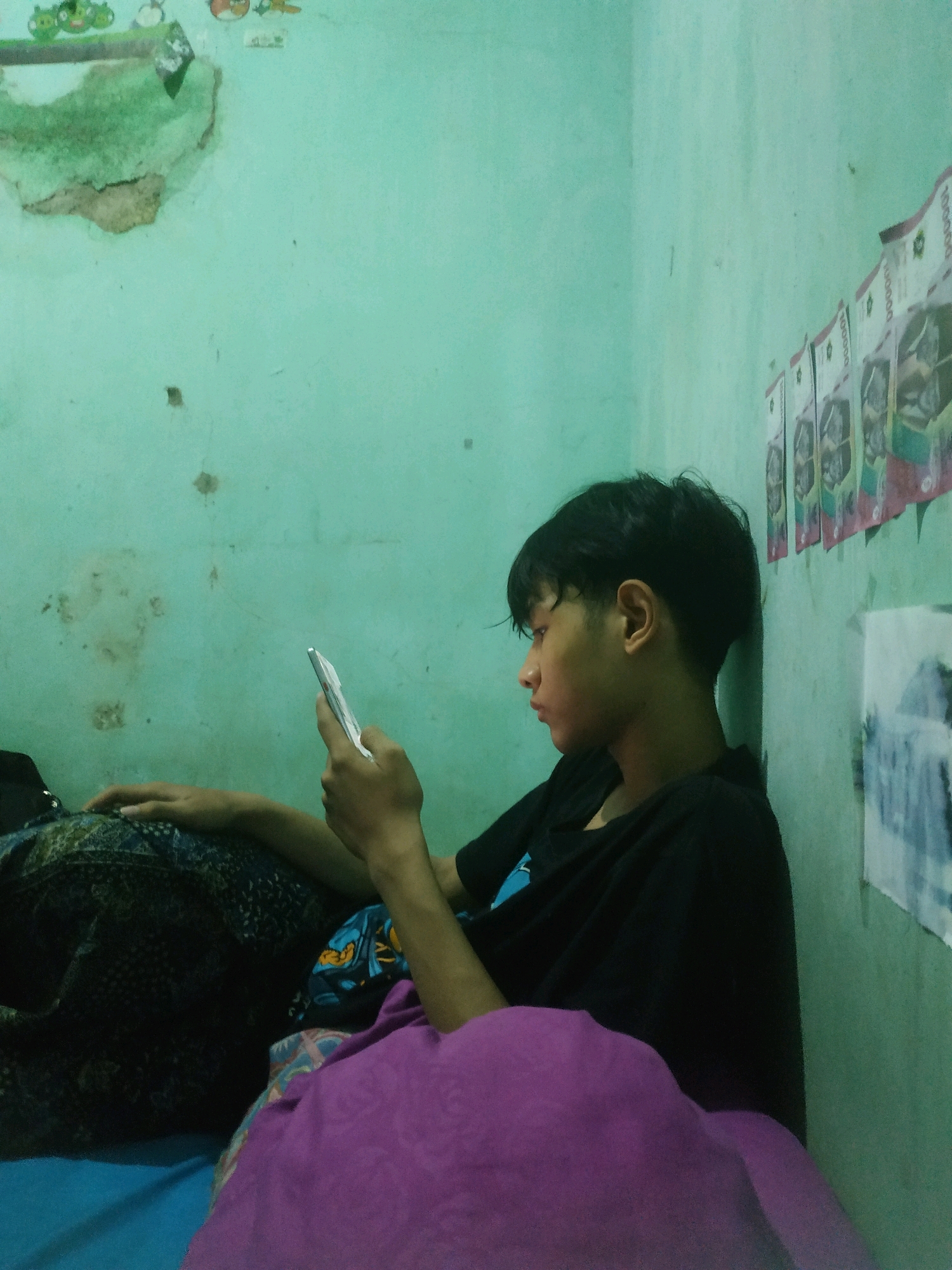 A young male with short, dark hair is leaning against a mint green wall in an indoor setting, possibly a bedroom. He is wearing a black t-shirt and is holding a smartphone in his right hand, appearing to be looking at the screen. He is partially covered by a patterned blanket in shades of dark blue and brown, and a purple blanket is in the foreground. The wall has some areas of wear and tear, with some peeling paint and discolored patches. There are also several pieces of paper with images of money affixed to the wall to the right of the male. The overall lighting is dim, suggesting it might be evening or the room is not well-lit. The scene is likely in Pacul, Indonesia, given the context provided.