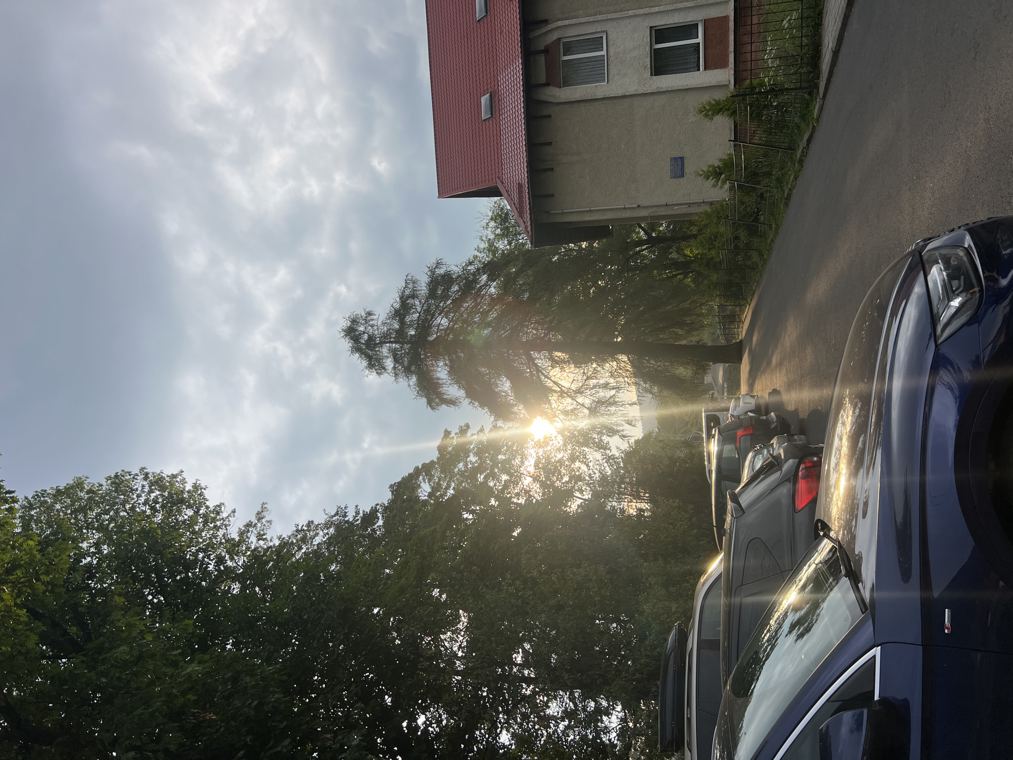 The image captures a street scene in Yaremche, Ukraine, likely in the morning or late afternoon due to the low angle of the sun casting long shadows. In the foreground, a dark blue car is parked on the left side of the asphalt road. Behind it, a line of other vehicles is visible, including SUVs and a car with a rooftop carrier. The road appears to be a typical residential or public street.
To the right of the road, a two-story building with a red tiled roof stands. The building's facade is a light beige color, and it features white-framed windows. A black metal fence runs along the front of the building, separating it from the road and a strip of greenery. Small, manicured evergreen trees are planted behind the fence.
Dominating the left side of the image and extending into the background are lush green trees, some of which are quite tall and dense. The sun is shining brightly through the foliage, creating lens flare and streaks of light across the scene. The sky is partly cloudy, with the sun's rays breaking through in some areas. The overall impression is a peaceful, somewhat overcast day with the sun intermittently breaking through the clouds, illuminating the parked cars and the building. There are no people visible in the image.