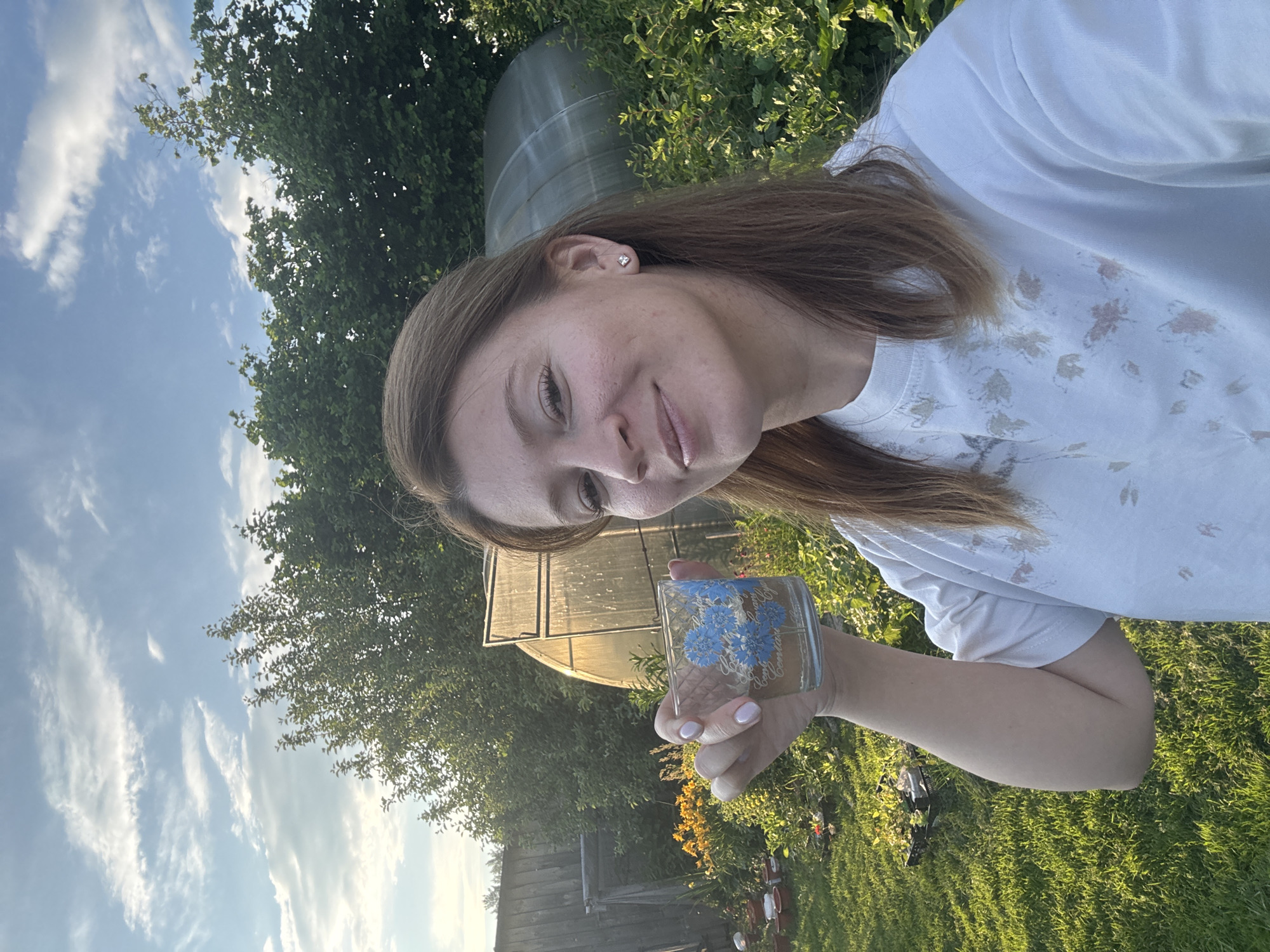 A woman is taking a selfie in a garden in Вассята, Russia. She is holding a glass with a blue floral pattern. She is wearing a white t-shirt with a floral print. In the background, there is a greenhouse, trees, and a wooden fence. The sky is partly cloudy.