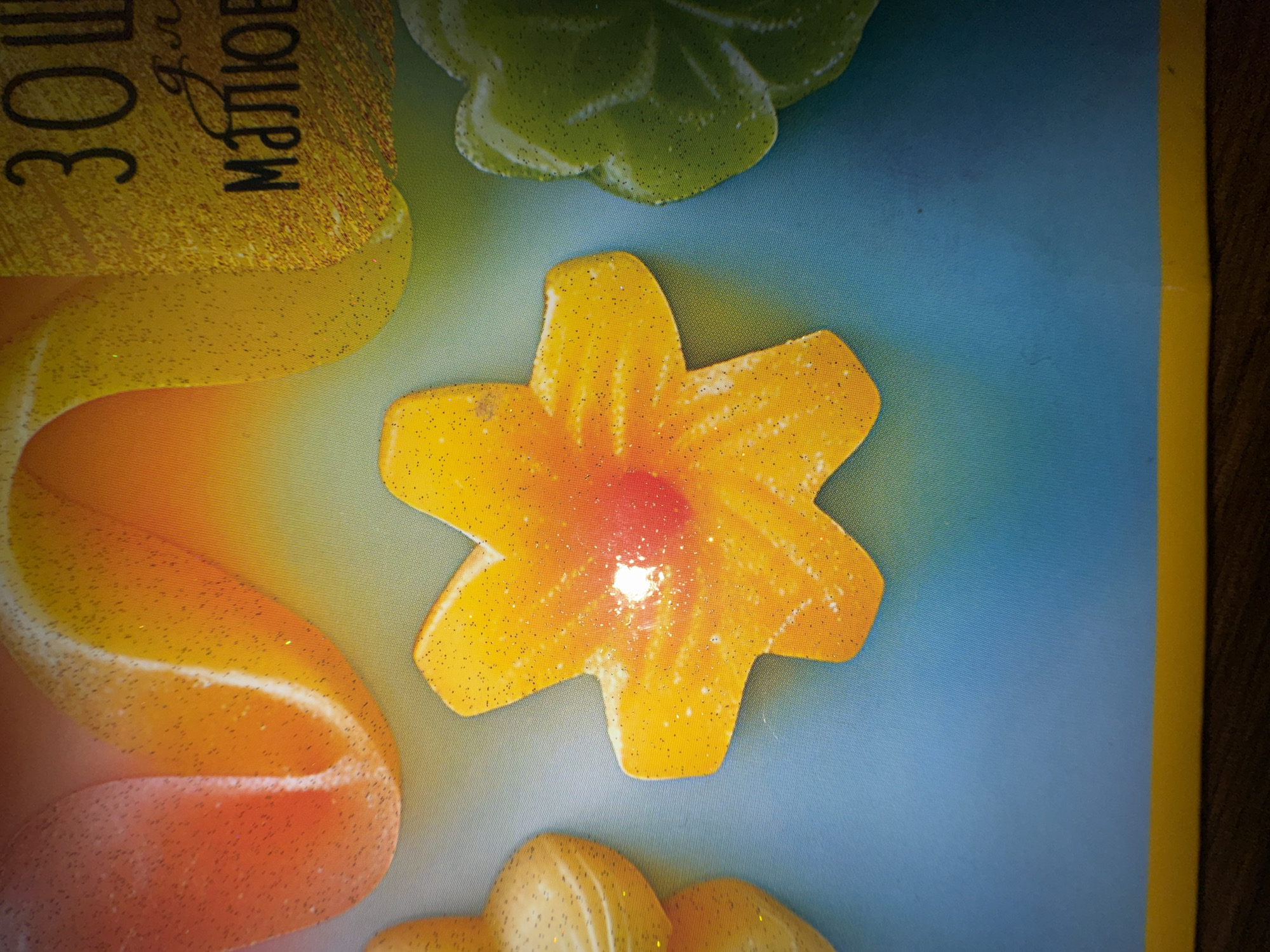 This is a close-up photograph of a colorful background with several glittery, candy-like objects resembling flowers and a ribbon. The objects are in various shades of orange, yellow, and green. There is text in Russian on the upper right side of the image. The background transitions from a light orange at the top to a bright blue at the bottom. A yellow border runs along the bottom of the image. The photograph does not depict any specific location in the city of Poltava, Ukraine, as it appears to be a product shot or a close-up of a decorative item. There are no people, actions, or interactions visible.