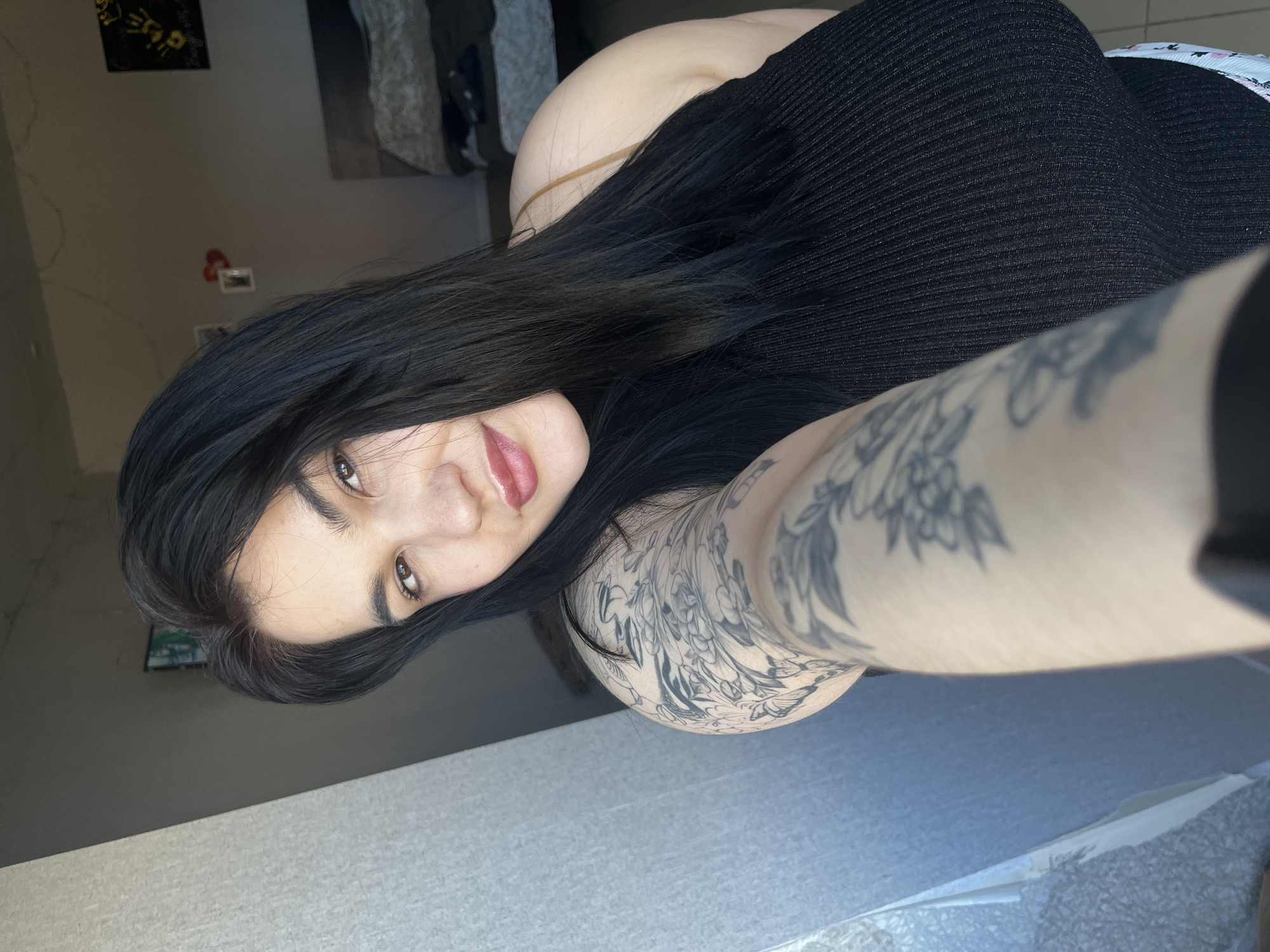 A selfie photograph shows a person with long, dark hair, fair skin, and full lips. They are wearing a black ribbed top. Their left arm is visible, covered in a large, detailed tattoo. The background is a blurry mix of interior elements, including a wall with framed artwork and a glimpse of what appears to be a bed. The lighting is soft and diffused. The location is not identifiable as Chaikovsky, Russia.