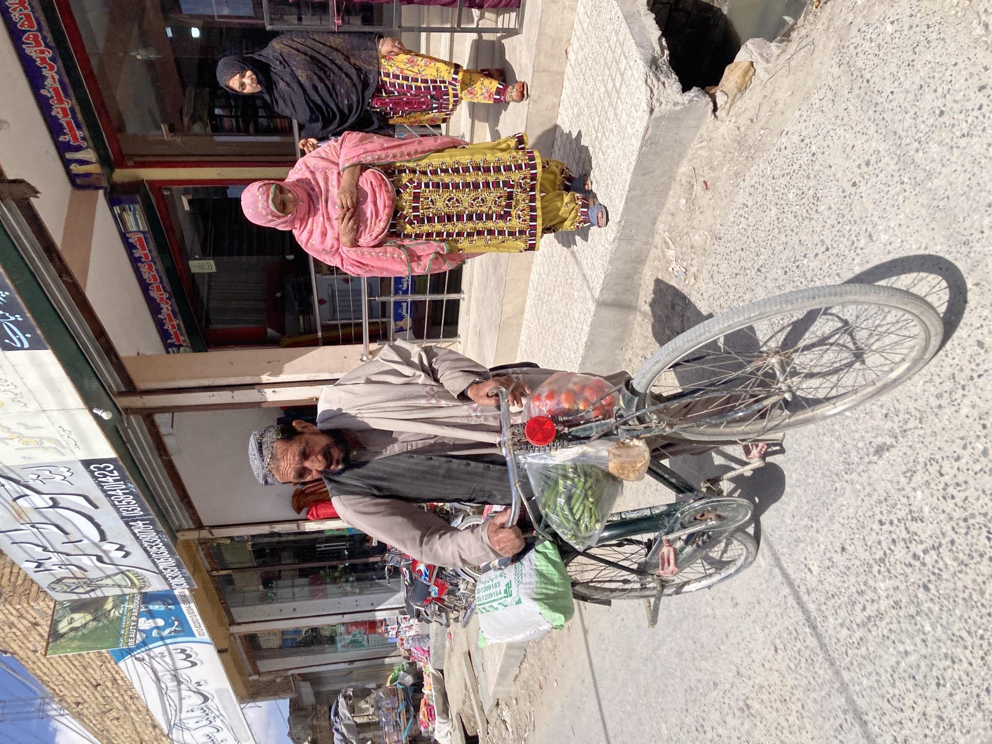 The image depicts a street scene in Quetta City Tehsil, Pakistan. An older man wearing a head covering and jacket is riding a bicycle. The bicycle carries bags of groceries. Two women are walking on the sidewalk; one is wearing a pink head covering and a patterned yellow and burgundy dress, and the other is wearing a black niqab and a similar yellow and burgundy dress. A shop is visible in the background with signs in Urdu. The sidewalk has a hole in it. Other motorbikes are visible. The scene appears to be during daylight.