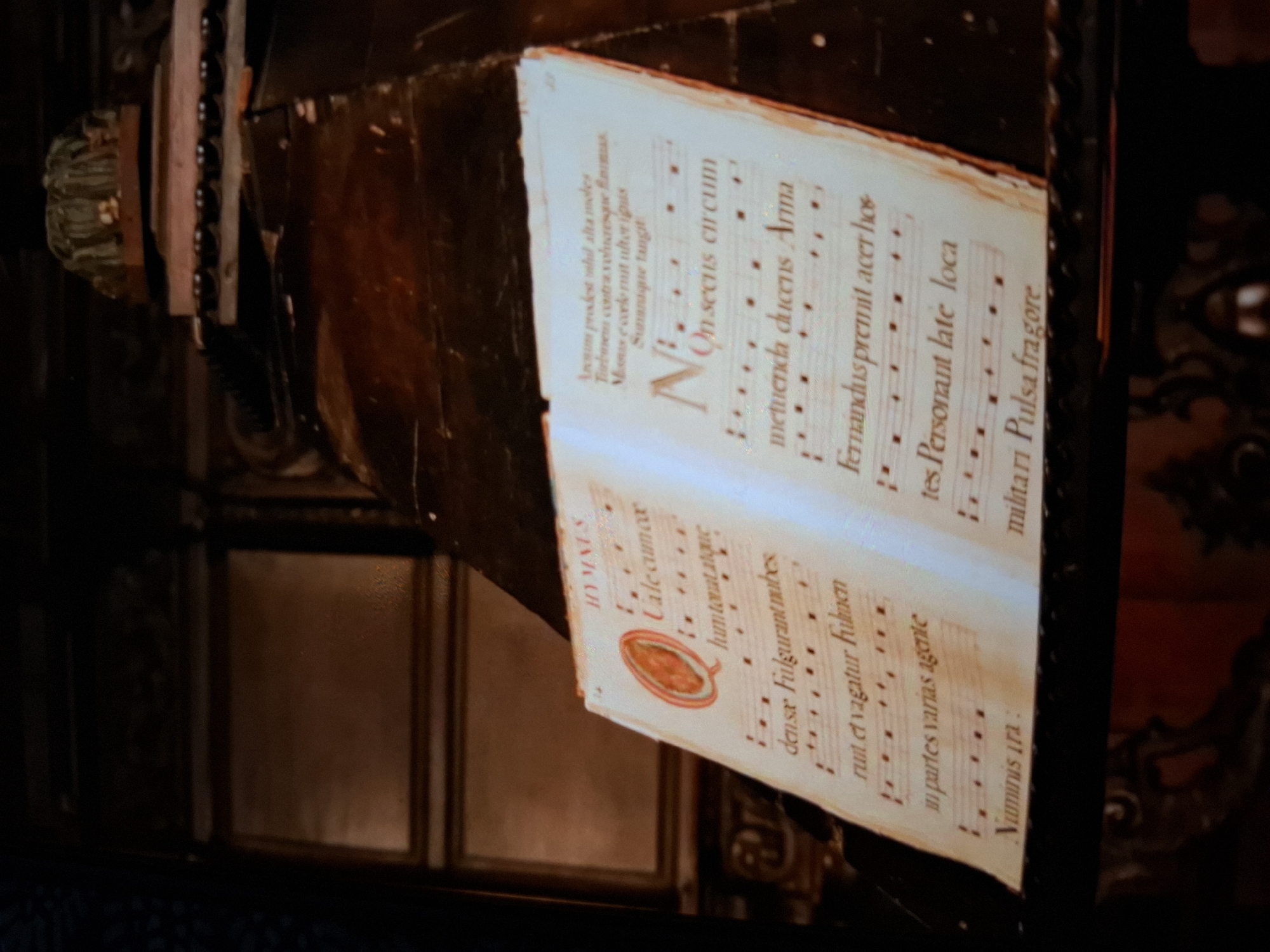 An old musical manuscript lies open on a dark wooden surface. The manuscript features text in Latin and musical notation. A large initial "Q" in red is on the left page. The right page has a large initial "N" in red. The background is dark and appears to be an interior, possibly a church or library, with ornate architectural elements visible. The overall lighting is dim, highlighting the manuscript.
