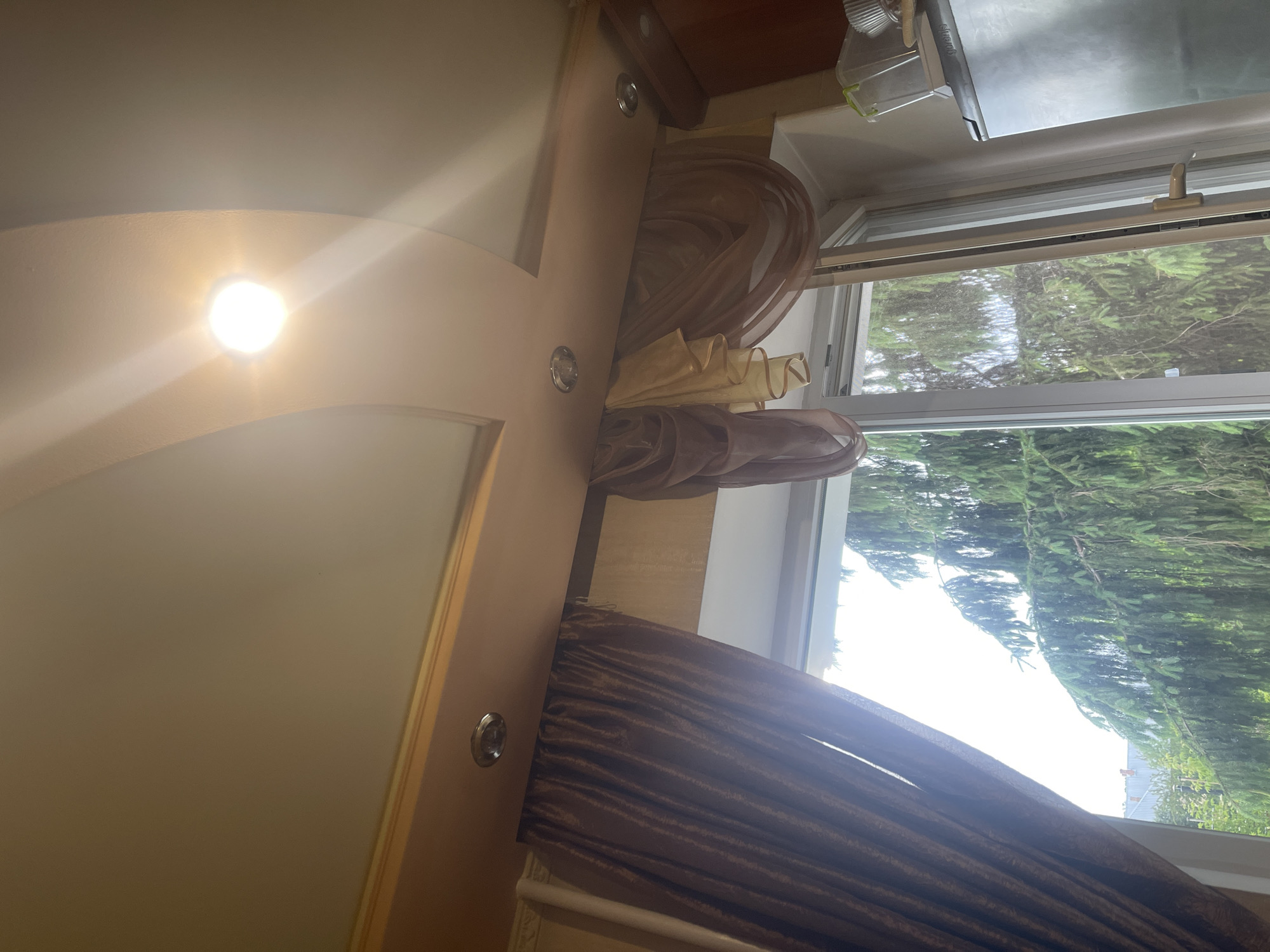 The image shows an interior view looking upwards and outwards through a window. The ceiling is adorned with recessed lighting and a decorative curved design. A valance with drapery, composed of thick brown curtains on the left and sheer, layered fabrics in shades of beige and purple on the right, hangs above the window. The window itself is open, revealing a dense canopy of green pine trees outside, with hints of rooftops visible in the background. To the right of the window, a portion of a stainless steel appliance, possibly a refrigerator, is visible, along with some containers. The scene appears to be a domestic interior looking out into a natural, possibly suburban or rural, setting.