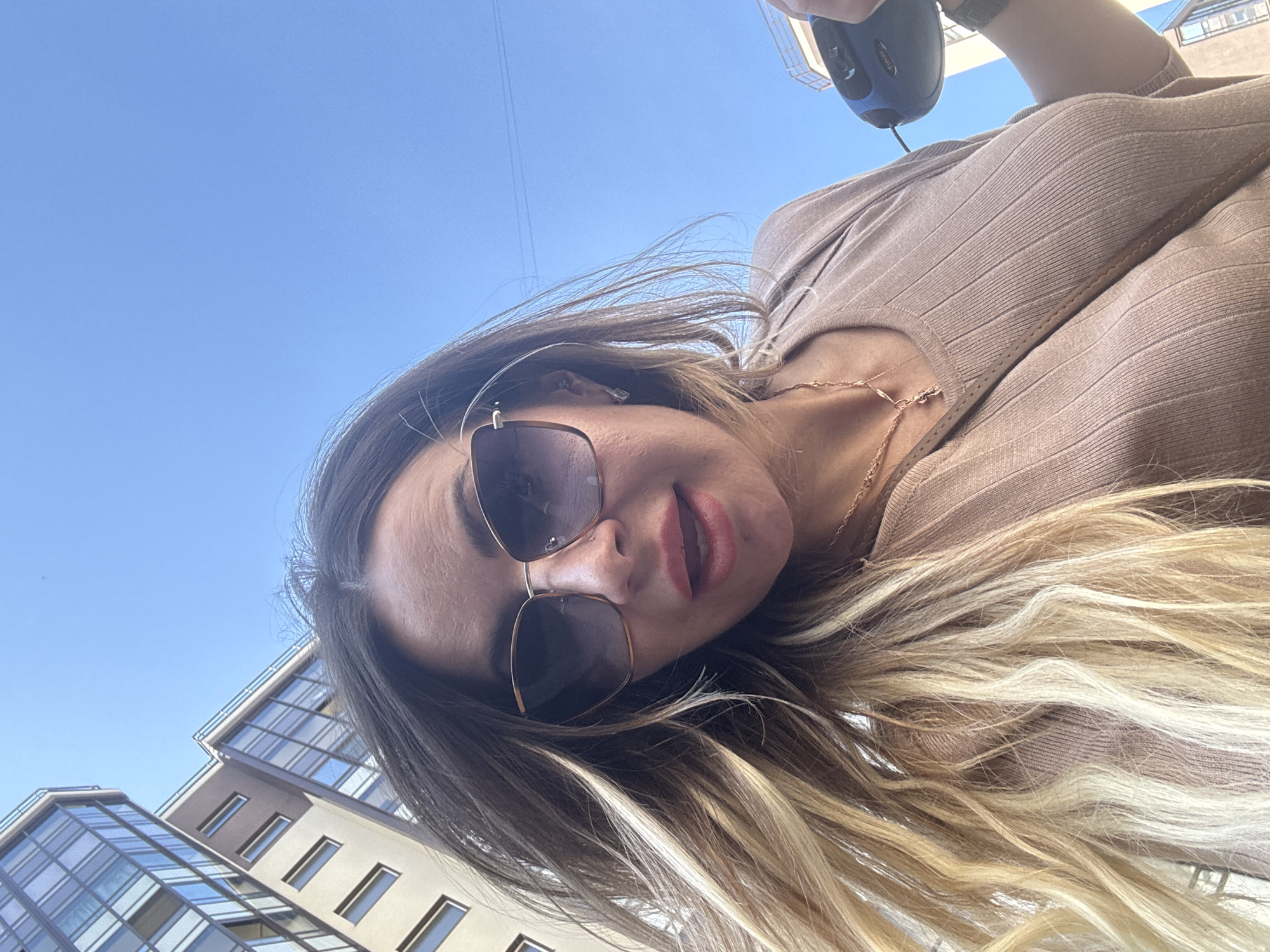 A low-angle, close-up selfie captures a person, likely female, outdoors against a bright blue sky and urban buildings. The subject's head is tilted, and she is looking directly into the camera with a slight smile or open mouth. She wears large, square-framed dark sunglasses, obscuring her eyes. Her long hair is styled with darker brown roots transitioning to blonde highlights and ends, flowing around her face and shoulders. She is dressed in a light brown, ribbed top, and a delicate gold-colored necklace is visible around her neck. In the upper right corner of the image, a dark blue and black handheld device with a cord can be seen, possibly being held by the subject.
The background features a clear, cloudless blue sky, indicating daytime and sunny weather. Below the horizon line, modern architectural structures are visible, characterized by multiple glass-paned facades and beige or brown wall sections, suggesting a contemporary urban environment. The buildings reflect the sunlight. The scene is set in Saint Petersburg, Russia. No discernible text is visible in the image.