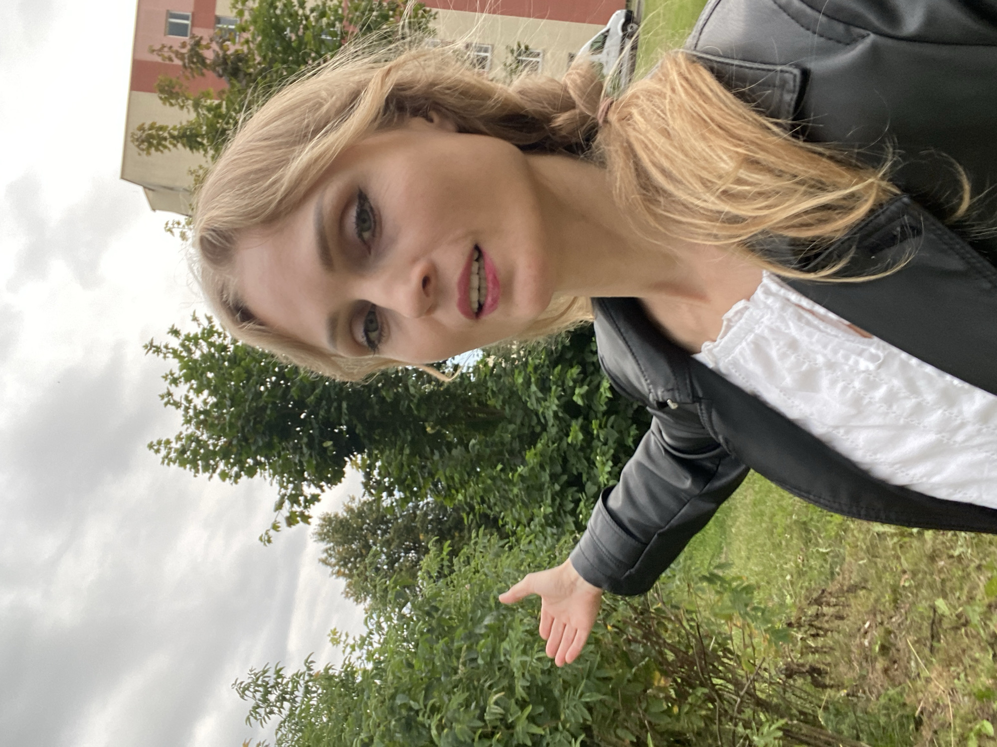 A woman with blonde hair in a braid is standing outdoors, gesturing to her left with her right hand. She is wearing a black leather jacket over a white top. Behind her, there are lush green trees, and in the background, a multi-story building with a pink and beige facade is visible. A white van is parked near the building. The sky is overcast. The scene appears to be in a residential area in the city of Lida, Belarus, possibly a courtyard or park.