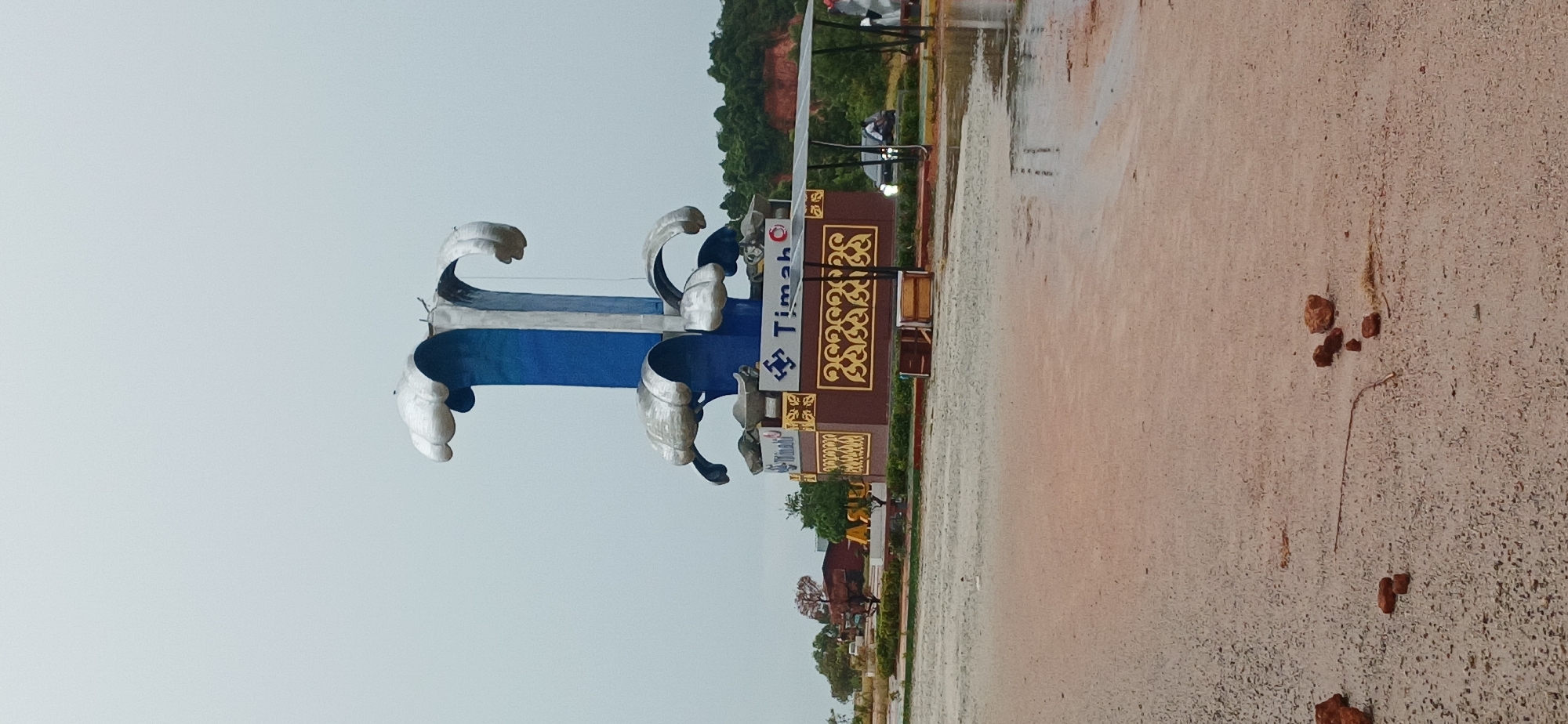 The image depicts a large, abstract sculpture in a public space, likely a roundabout or park, in Tanjung Balai Karimun, Indonesia. The sculpture is predominantly blue and silver, with wave-like elements at the top and a tiered base. The base is brown with gold decorative patterns and signage that includes the logo "Timah". In the background, there are trees, buildings, and a hilly landscape under an overcast sky. The foreground shows a wet, sandy or dirt surface, indicating recent rain or a water feature. Several scattered bricks and a thin stick are visible on the ground in the immediate foreground. A car is partially visible on the right side of the image, and another vehicle is further in the background.