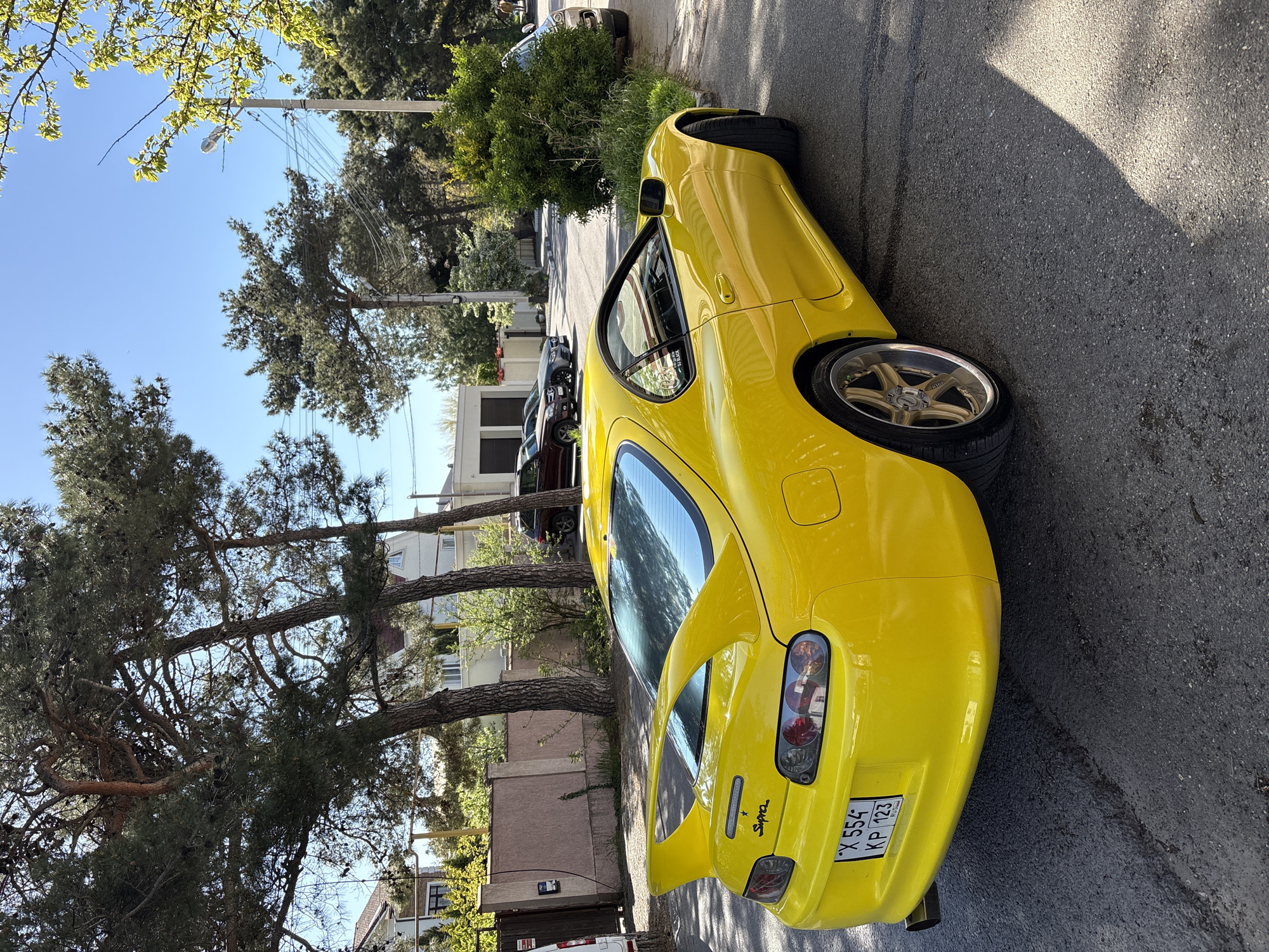 The image, rotated 90 degrees clockwise, depicts a bright yellow Toyota Supra (likely an MK4 model) parked on the side of an asphalt street in Gelendzhik, Russia. The car is angled with its rear facing towards the viewer, highlighting its iconic taillights and integrated spoiler. The glossy finish of the paint reflects the bright sunlight.
On the rear of the vehicle, the word "Supra" is visible, and the license plate displays "X 554 KP 123". The "123" regional code indicates Krasnodar Krai, consistent with the provided location of Gelendzhik. A decal partially visible on the side window reads "J. BLACK". The car is fitted with gold-colored multi-spoke wheels.
The setting is outdoors on a clear, sunny day, evidenced by the bright blue sky and the strong shadows cast on the asphalt road from the car and nearby objects. In the background, other cars are parked along the street, including a dark SUV or truck and a lighter sedan. Multi-story buildings, characteristic of a residential area, are visible beyond the street. Large, mature evergreen trees, likely pines, line the street, with their branches filling much of the background and upper frame. Power lines and a street light are also visible against the sky. No people are present in the image, suggesting a quiet, daytime street scene.