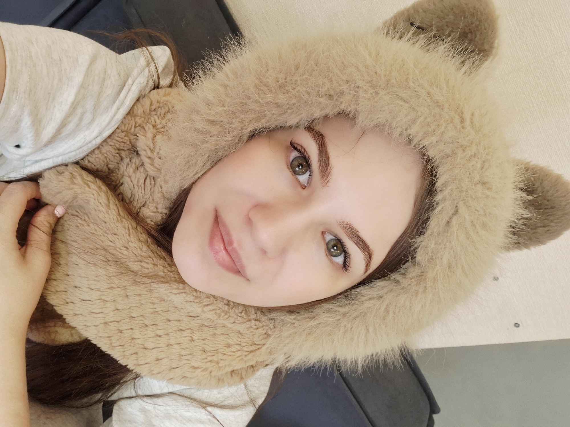 A young woman is prominently featured in a close-up portrait, lying down or reclining indoors and looking directly at the camera with a gentle, inviting expression. She has long, dark brown hair peeking out from under a light brown faux fur hood, which has distinct, upright ear-like structures, giving it an animalistic, possibly bear or cat, appearance. Her eyes are a striking green or hazel, complemented by natural-looking makeup, including well-defined eyebrows and curled eyelashes. Her lips are a soft, natural pink and slightly parted in a subtle smile.
She is wearing a light-colored, possibly off-white or beige, top. Her left hand, adorned with light pink nail polish, rests softly on the faux fur of the hood. The background includes dark fabric, likely a sofa or chair on which she is resting, and a plain, lighter-colored surface to her right. The overall mood of the image is cozy and relaxed. No specific activity is depicted other than posing for the photograph. There are no discernible location cues within the frame to indicate Chaikovsky, Russia, nor is there any visible text. The lighting suggests an indoor setting, likely during the day or a well-lit evening.