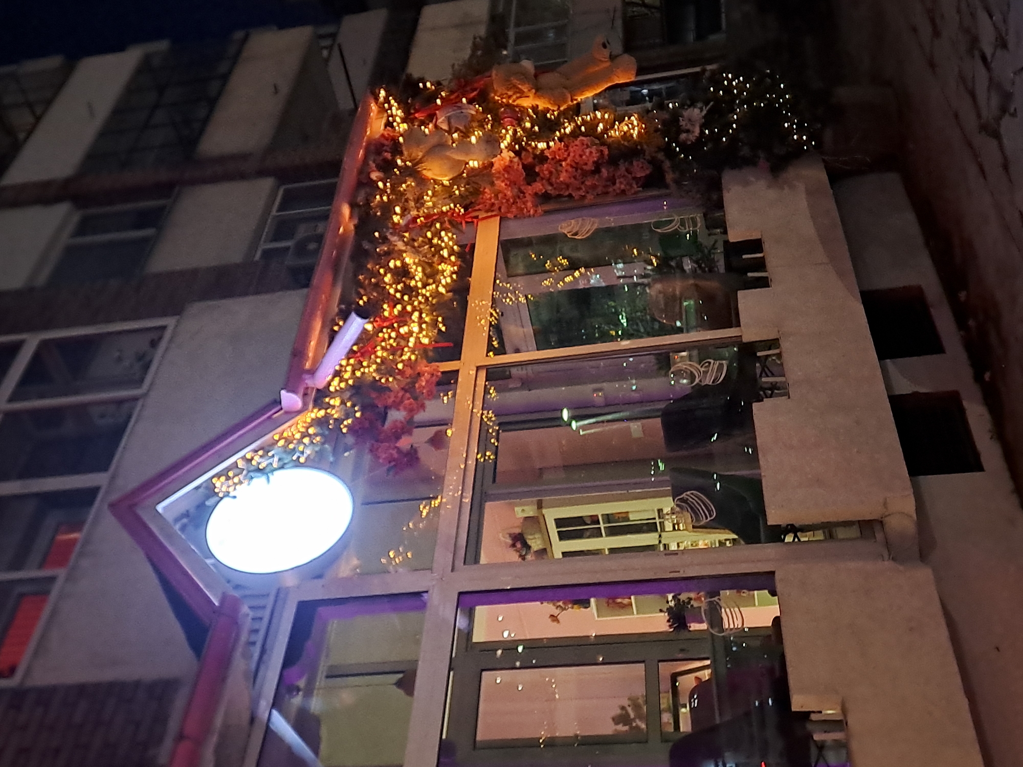 The image shows a building in Veliko Tarnovo, Bulgaria, decorated with lights and ornaments. The building appears to be a business, as it has a circular sign with a blank face at the entrance. The facade is modern, with large windows. The building is multi-storied, and other buildings can be seen in the background. The lighting indicates that it is nighttime. Ornaments are placed on the roof and around the sign.