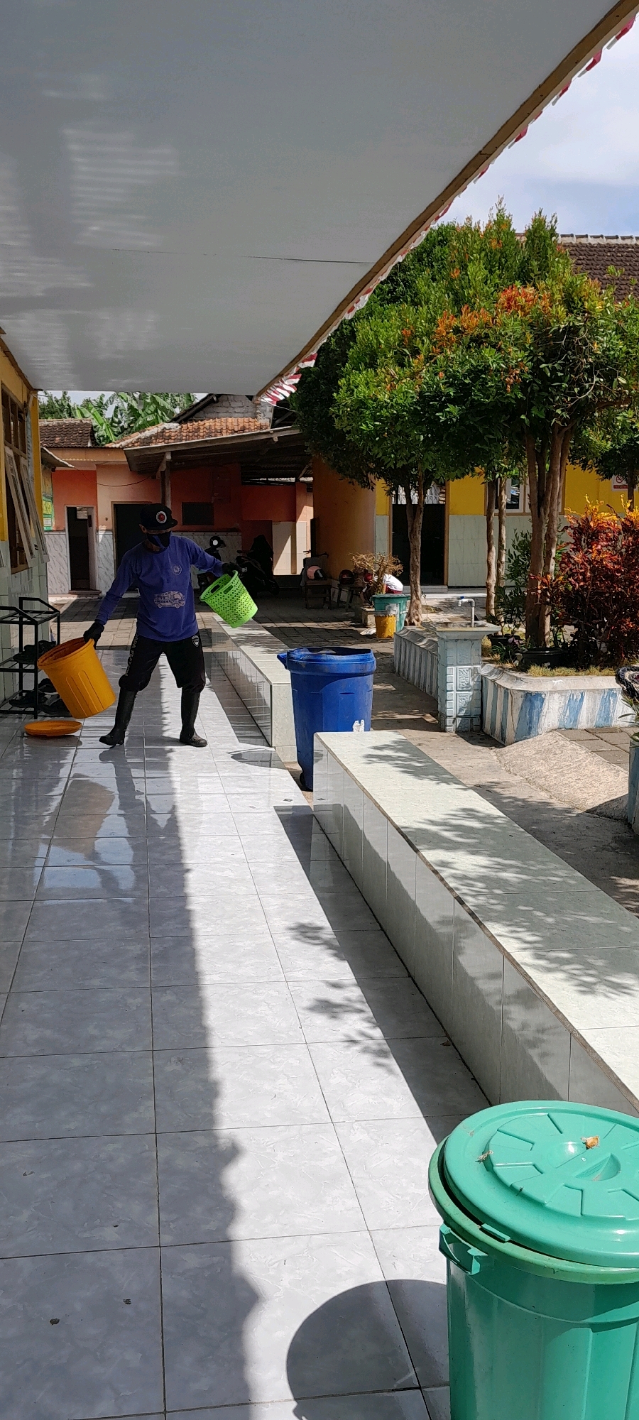 A man wearing a blue long-sleeved shirt, black shorts, black boots, a black baseball cap, and a black face mask is standing on a tiled walkway. He is holding a yellow bucket in his left hand and a green basket in his right hand. The man appears to be in motion, possibly cleaning or carrying items. The walkway is covered by a white awning. To the right of the man, there is a blue trash can and a low, tiled bench. In the background, there are trees with green and orange foliage, buildings with orange and yellow walls, and a motorcycle. The scene is outdoors, illuminated by sunlight casting shadows on the tiles. The location is identified as Rogojampi, Indonesia.