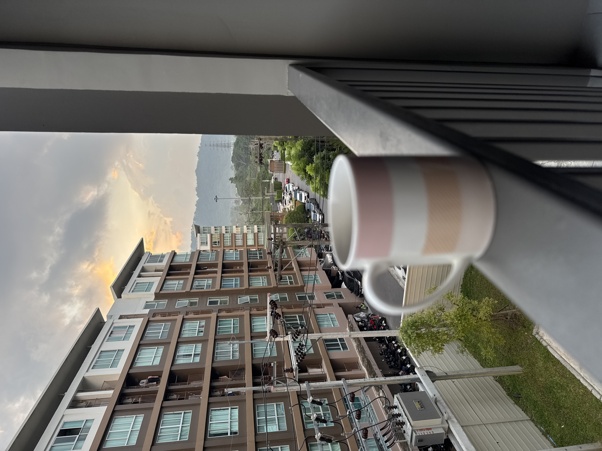 A mug with pink and cream stripes sits in the foreground on a balcony railing, partially obscuring a view of Kathu, Thailand. In the mid-ground, a multi-story apartment building with brown and white facades dominates the left side of the frame, with balconies visible on each floor. The building is adjacent to a street lined with parked motorcycles and cars. Further in the background, a road curves towards a landscape of green hills and trees under a cloudy sky with hints of orange and yellow from a setting or rising sun. Electrical poles and wires are visible throughout the scene.