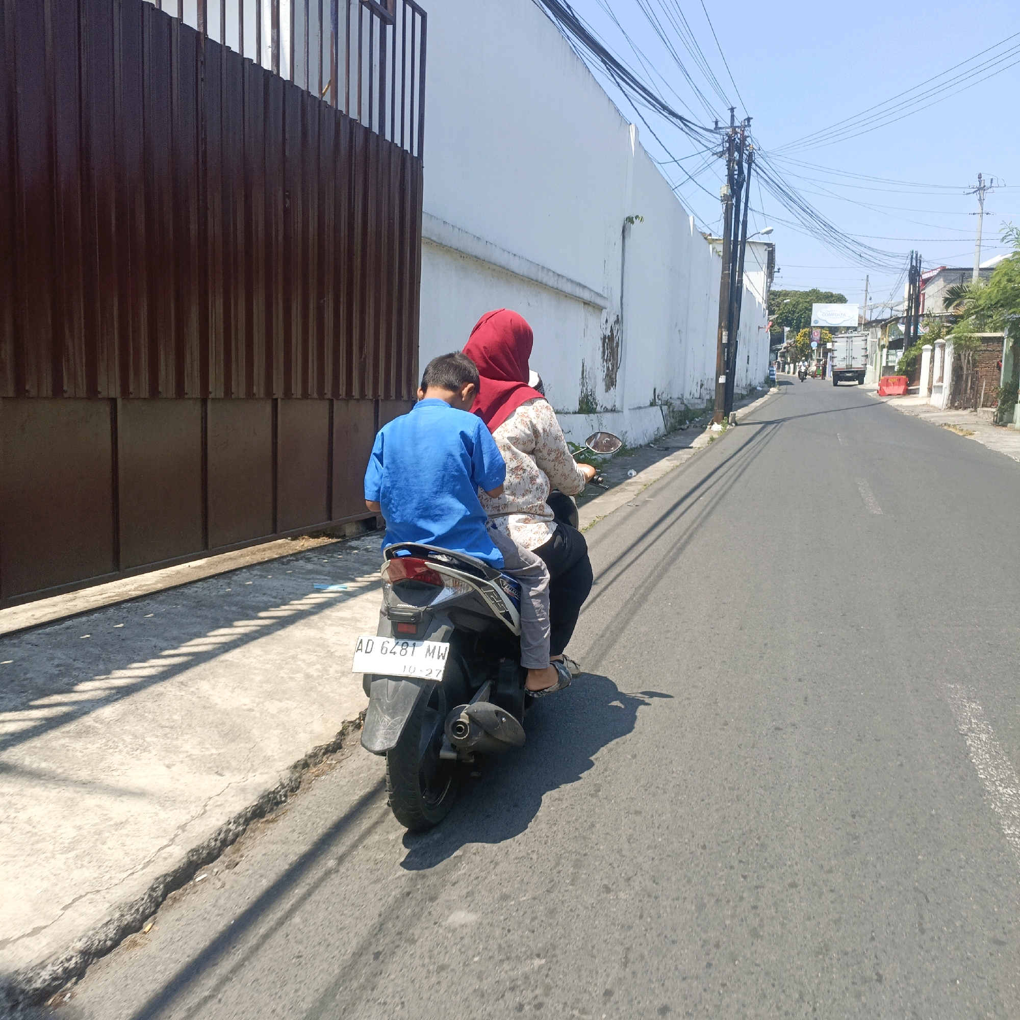 This is a medium shot, taken from behind a motorbike, on a sunny day in Salatiga, Indonesia. The motorbike is traveling on an asphalt road, with a concrete sidewalk and a dark brown corrugated metal fence on the left. A woman wearing a red hijab and a patterned shirt is driving the motorbike, with a young boy wearing a bright blue shirt sitting behind her. The motorbike has a white license plate that reads "AD 6481 MW" and "10-27". In the background, there is a white building, electrical poles with many wires, and other buildings and trees lining the street. The scene depicts everyday life in a city.