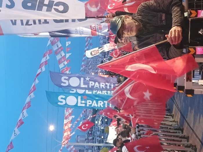 The image shows a gathering of people in a public space, most likely a political rally or demonstration. The event is held in Istanbul, Turkey at the coordinates 41.084 latitude and 28.678 longitude. The image depicts a large group of people holding flags. Several people are visible in the foreground, many wearing red and white colors. Several flags are hanging across the image, including Turkish flags and banners with the text "SOL PARTI" and "CHP." The foreground subject is a middle-aged man wearing a black jacket and glasses, holding a Turkish flag. He appears to be talking with someone to the right of the frame. In the background, there are numerous other people, most of them holding red and white flags and banners, and a few with the CHP flags. The environment is likely a city park or a public square. The scene is dominated by the flags and the crowd, with the cityscape in the background. The lighting is relatively low, suggesting that the image was taken in the late afternoon or early evening.