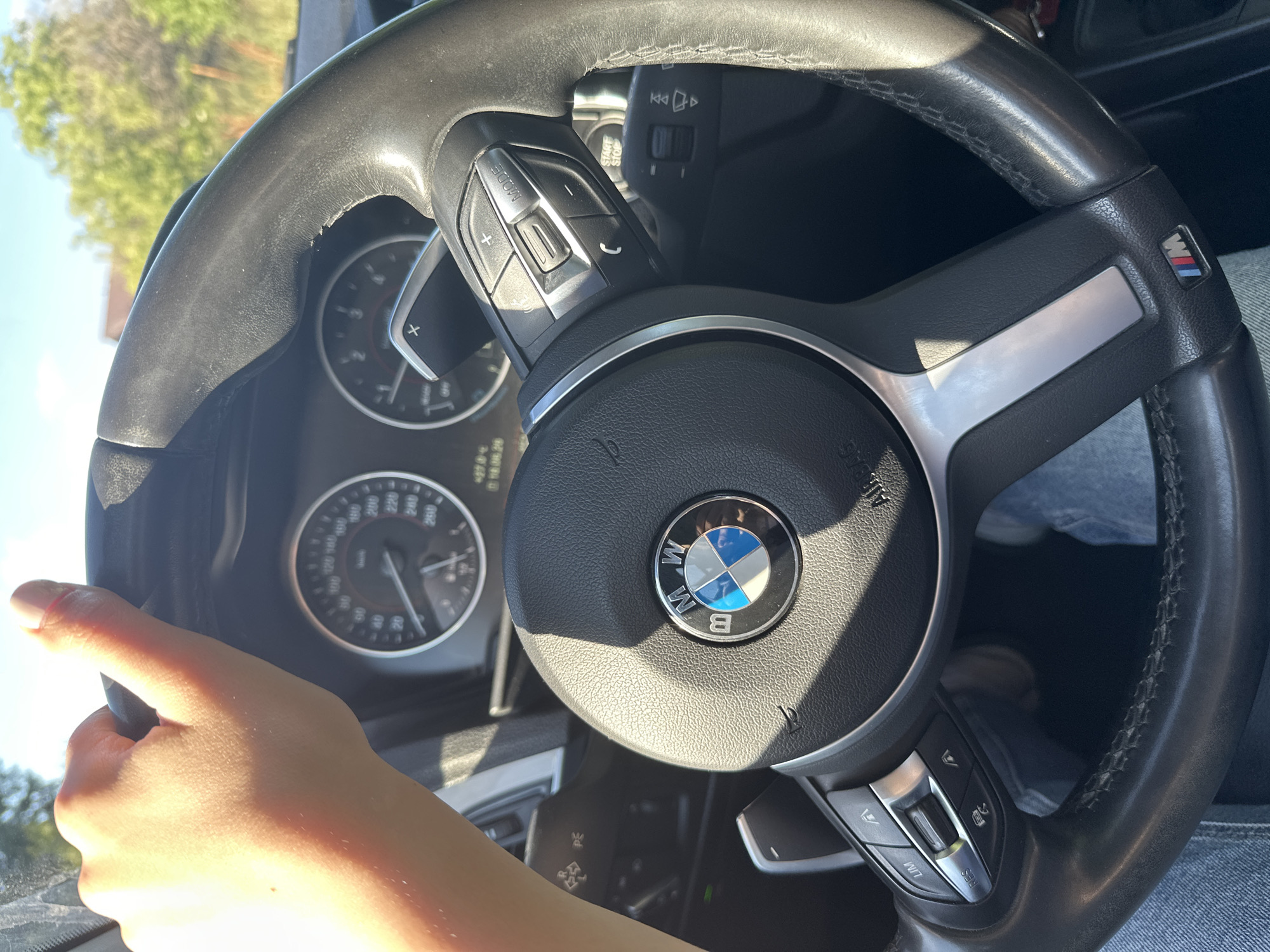 The image shows a close-up of a person's hands on the steering wheel of a car, as seen from the driver's perspective. The steering wheel is black leather with silver accents and features the BMW logo in the center. On the left side of the steering wheel, there are control buttons for volume and mode, and on the right, there are buttons for cruise control and limiter functions. The car's dashboard instruments, including a speedometer, are visible behind the steering wheel, with the needle on the speedometer indicating a speed of approximately 120 km/h. The sunlight suggests it is daytime, and the clear sky implies good weather. The background is blurred, but hints of green trees and a bright sky suggest an outdoor setting, likely a road. The overall impression is of someone driving on a sunny day. Visible text includes "BMW" on the steering wheel and "M" logo on the steering wheel. There is also some text on the dashboard instruments which is not fully legible. The scene is set in Gorni Bogrov, Bulgaria.