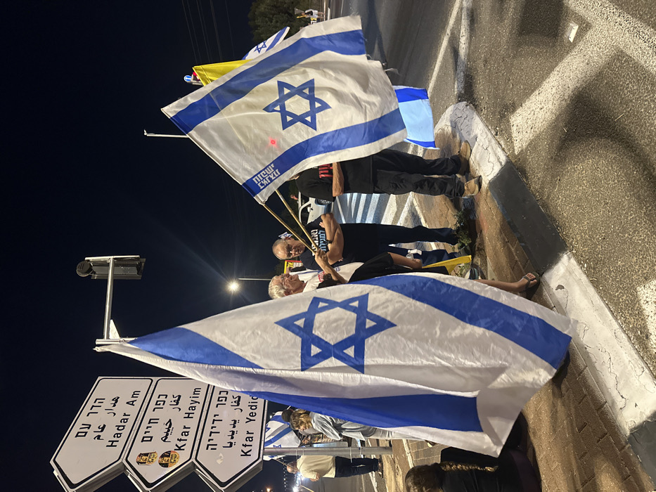 The image captures a nighttime gathering on a brick-paved sidewalk bordering an asphalt road, illuminated by artificial streetlights. The location is an intersection in the vicinity of Mishmar Hasharon, Israel, indicated by a prominent street sign displaying directions to "Hadar Am," "Kfar Hayim," and "Kfar Yedidia" in both Hebrew and English. A traffic light signal is mounted above the signs.
Approximately eight to ten individuals are visible, predominantly adults. Multiple Israeli flags are held by the participants; these flags feature a blue Star of David on a white field, bordered by two horizontal blue stripes. At least one flag prominently displays the Hebrew text "מגינים למדינה" (Defending the State). Three men are central to the foreground, appearing to converse while holding flags. The man on the left wears a light-colored shirt and glasses. The man in the center wears a dark t-shirt with his arms crossed, also holding a flag. The man on the right, partially obscured by a flag, wears a dark t-shirt and dark pants. A person is seated on the curb in the lower-left foreground. In the background, a white SUV and other vehicle lights are discernible on the road. The road features white painted markings.