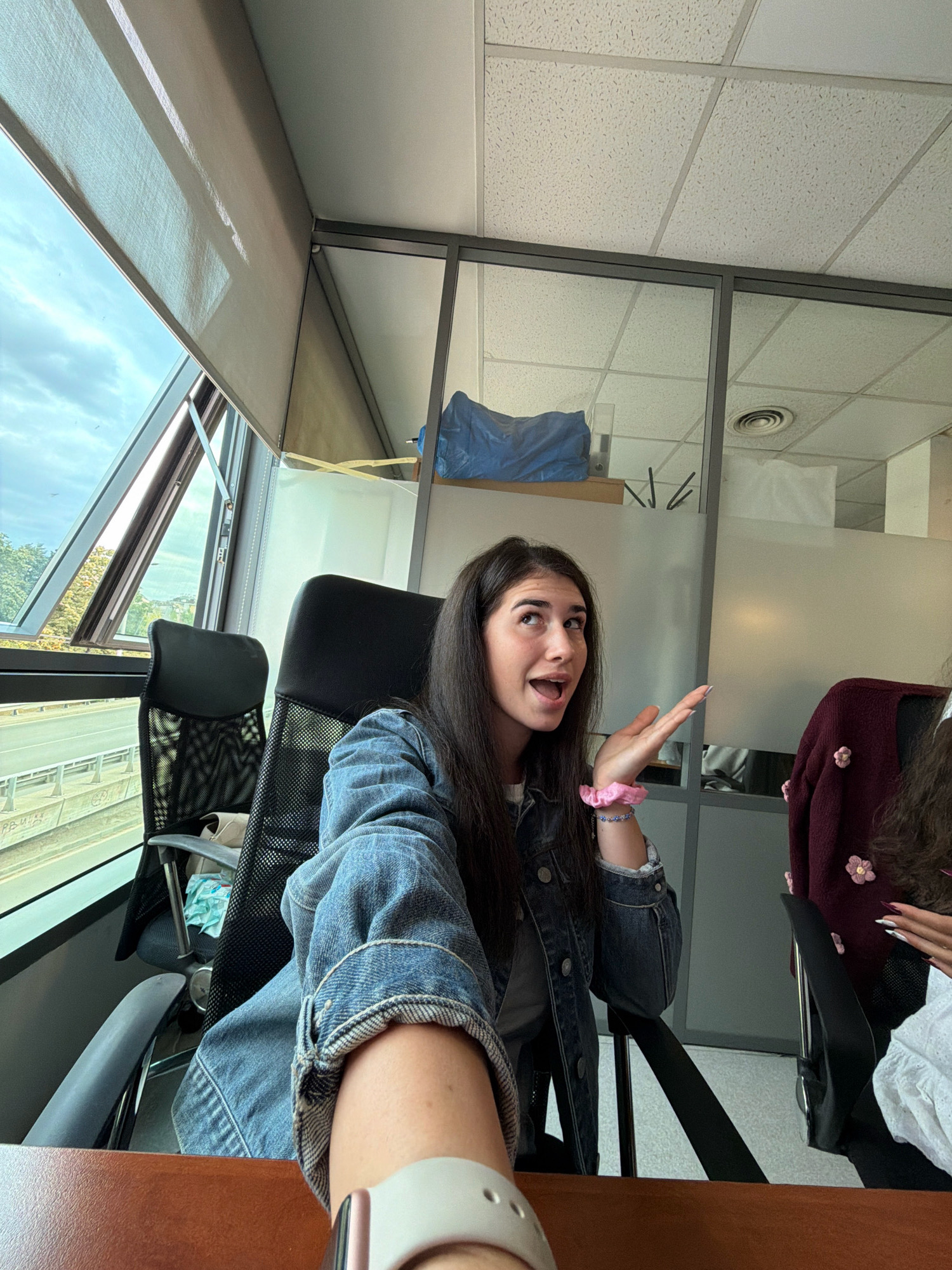 A young woman with long dark hair, wearing a denim jacket and a pink scrunchie on her wrist, is seated in an office chair, looking upwards and to her left with her mouth open as if speaking. She is in an office environment with large windows showing a cloudy sky and some greenery outside, suggesting a location in Sofia, Bulgaria. The office has glass partitions, a suspended ceiling with acoustic tiles, and other chairs are visible. A blue bag is on a shelf in the background. The woman's left arm is extended towards the camera, holding what appears to be a phone taking a selfie. Another person's shoulder and arm, wearing a burgundy sweater with floral appliques, are visible to the right.