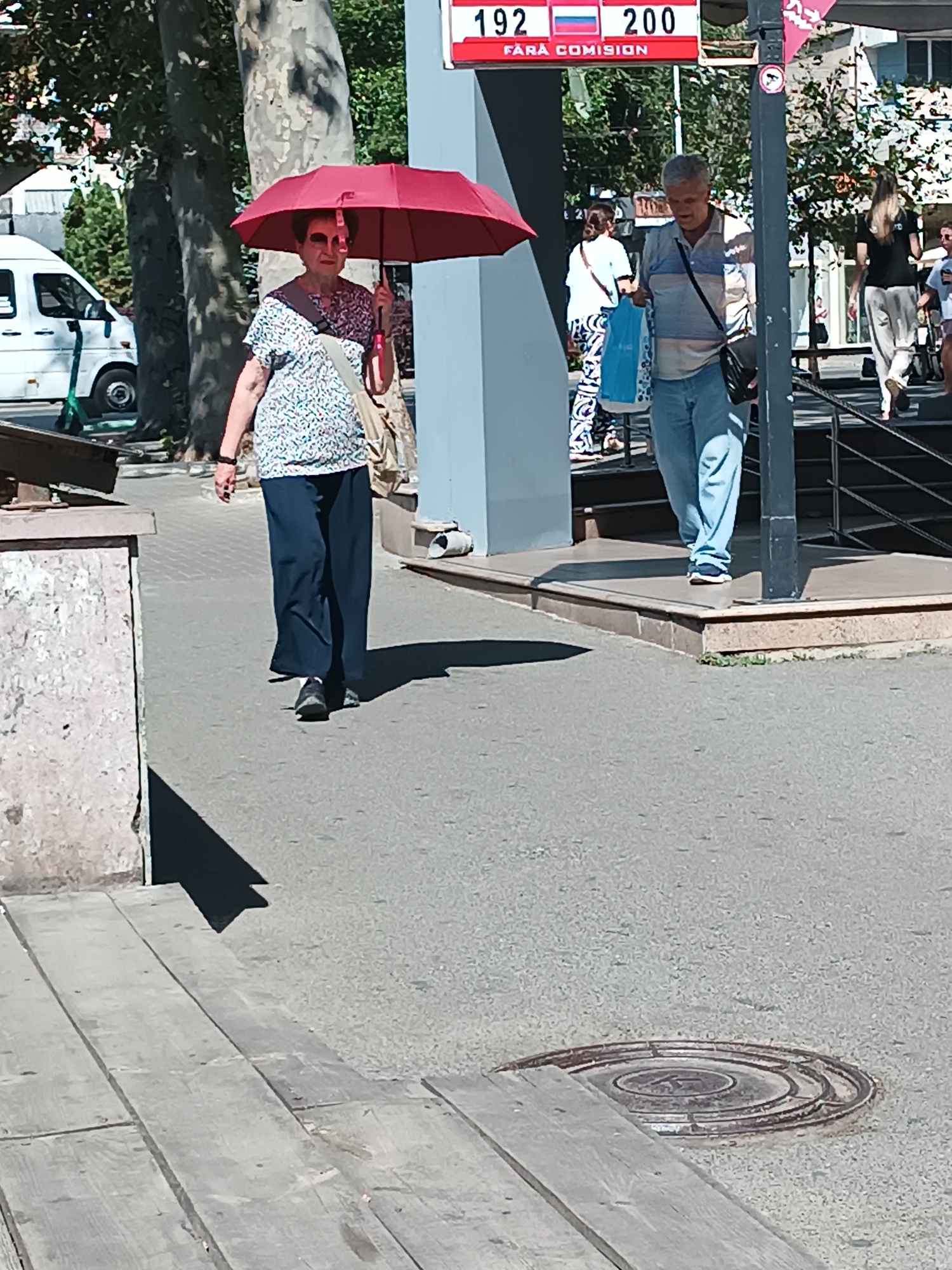 The image depicts a street scene in Chișinău, Moldova, likely on a sunny day, as indicated by the presence of an umbrella and shadows. In the foreground, a woman with short dark hair walks from left to right, holding a red umbrella over her head. She is wearing a patterned blouse and dark, loose-fitting pants. To her right, a man walks in the same direction. He has short grey hair and is wearing a striped polo shirt and light blue jeans. He is carrying a dark bag on his shoulder and a blue shopping bag in his left hand. Behind them, other people and elements of urban infrastructure are visible, including a white van, trees, a sign with red and white text, and a metal railing. A circular manhole cover is visible in the pavement in the lower right portion of the image. The overall impression is of an everyday street scene in a city.