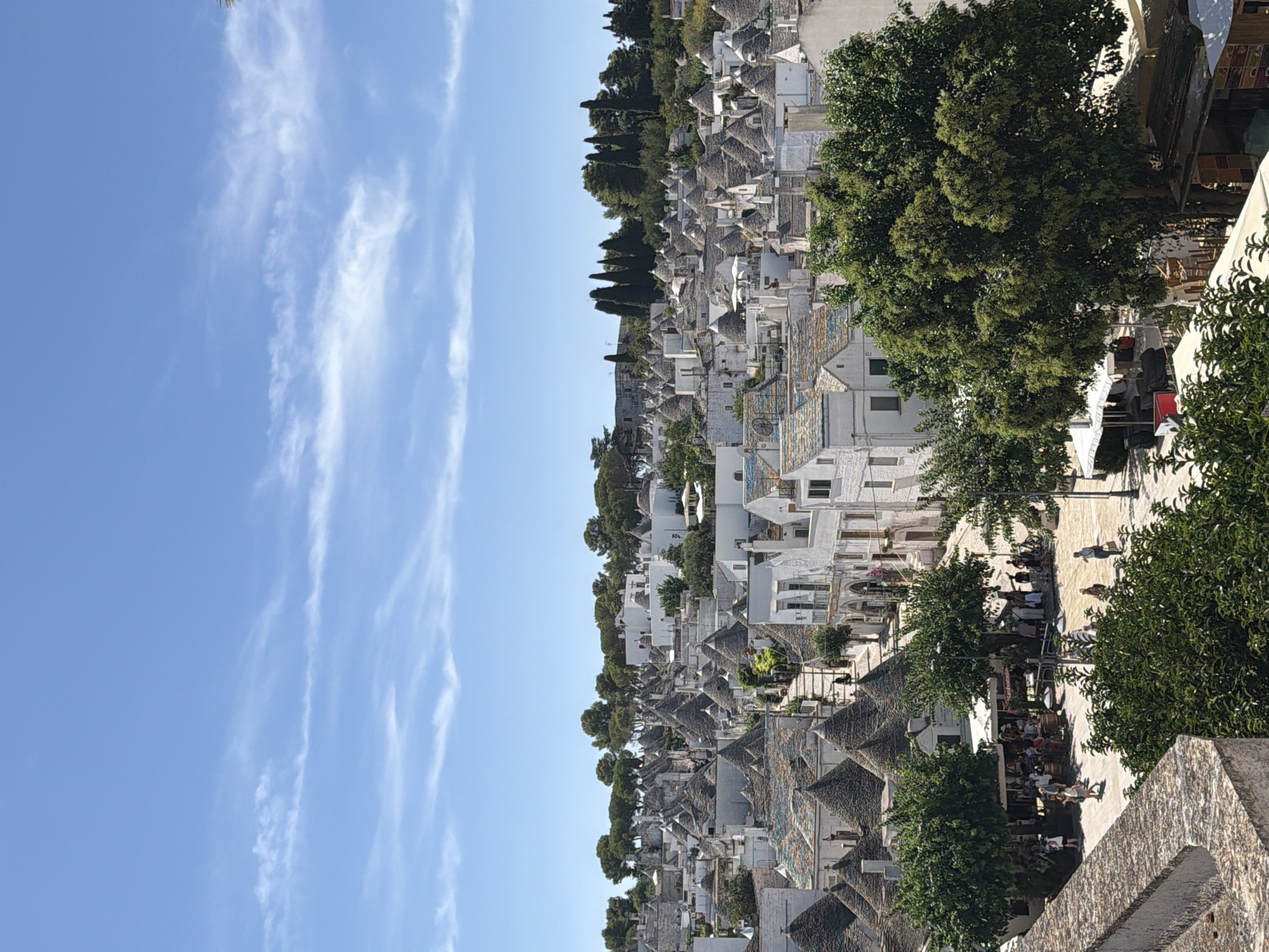 The image depicts a high-angle view of Alberobello, Italy, showcasing its iconic trulli houses under a bright blue sky with scattered white clouds. The foreground is dominated by a stone wall and lush green trees, with a view extending over a dense cluster of white trulli buildings with conical stone roofs. A pathway winds through the village, lined with more trulli and a few trees. In the mid-ground, a small square or gathering area is visible with people walking, browsing stalls under awnings, and what appear to be cafe tables and chairs. Further into the distance, the trulli extend up a hillside, interspersed with evergreen trees. The architecture is predominantly white with grey stone roofs, some of which are adorned with colorful patterns. The overall impression is of a sun-drenched, historic European town.
