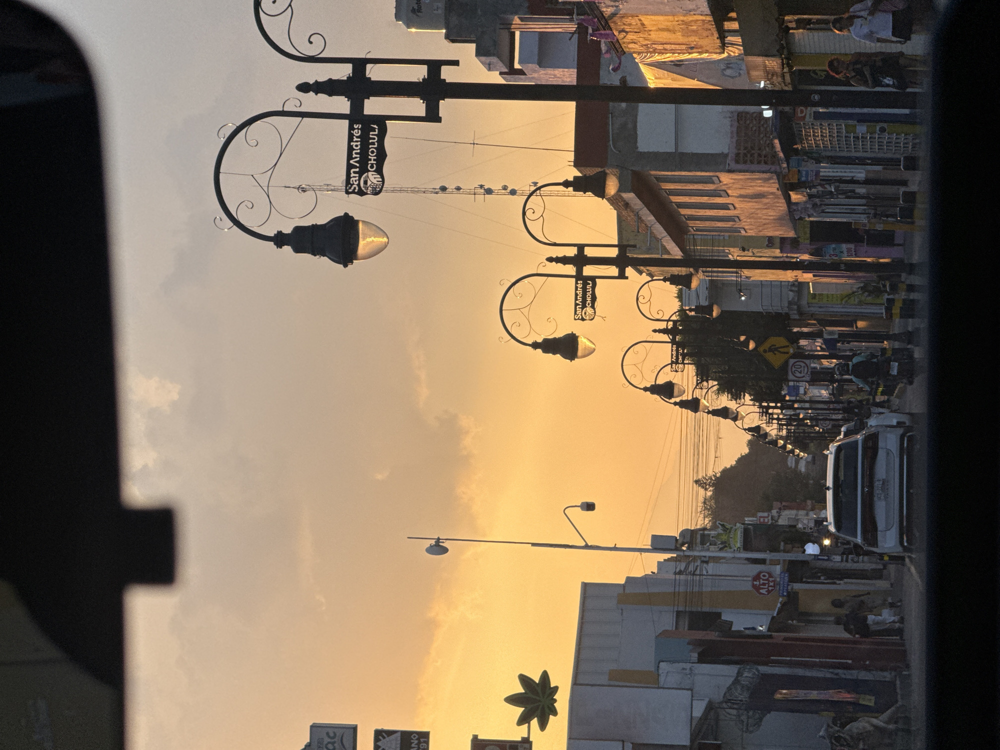 Here is a concise description of the image:
The image captures a street scene in San Andrés Cholula at sunset. Rows of ornate lampposts with "San Andrés CHOLULA" signs line the street. Buildings of various heights and architectural styles flank the road. A few people are visible on the sidewalks, and vehicles including a white truck are moving down the street. The sky is a gradient of orange and yellow. The image is taken from inside a vehicle.