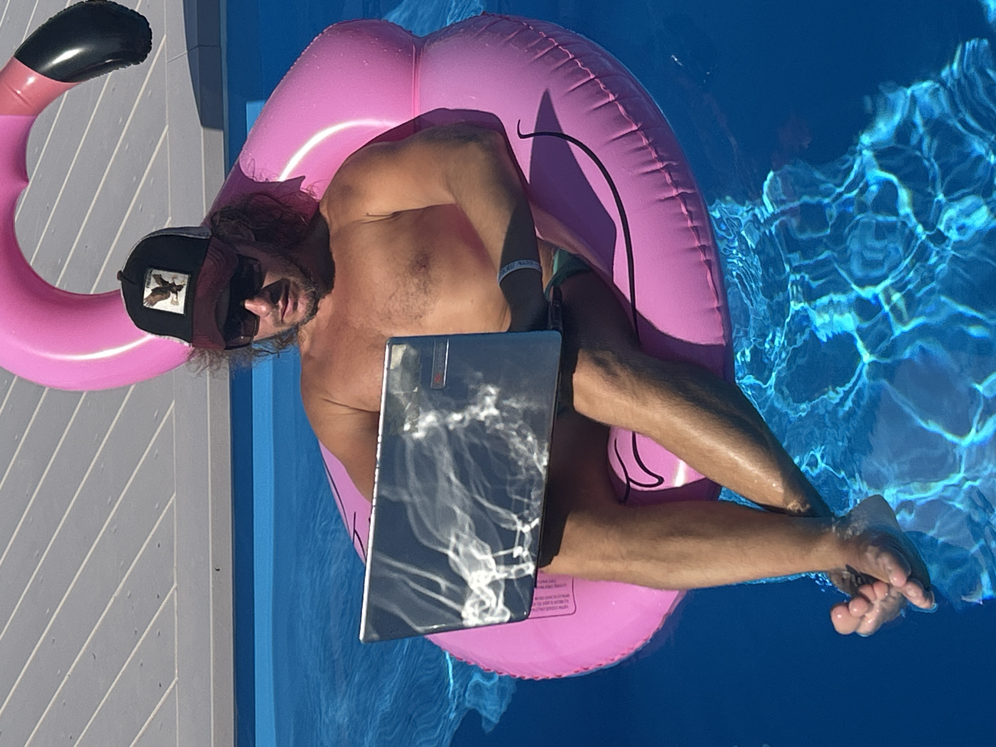 A shirtless man wearing a baseball cap and sunglasses is sitting on a pink inflatable flamingo in a swimming pool. He is holding a silver laptop on his lap and appears to be working. The swimming pool is surrounded by a wooden deck. The man has long, curly brown hair and is wearing swim trunks. The water in the pool is blue and clear, with sunlight reflecting on the surface. The man has a bracelet on his left wrist. The scene takes place outdoors on a sunny day.