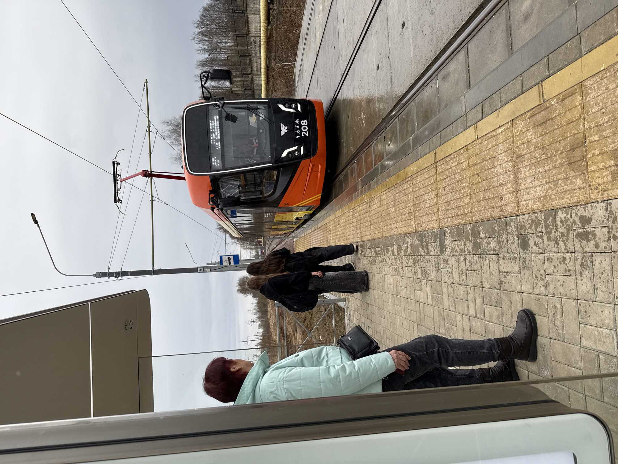 The image depicts a streetcar at a station in Unknown, Unknown. Three individuals are visible on the platform: one wearing a light blue jacket and dark pants, and two younger figures in dark clothing. The streetcar is orange and grey with the number "208" displayed on its front. The platform is paved with yellow tactile paving along the edge. A grey sky is overhead. The streetcar is adjacent to the platform with visible tracks, and a lamp post. Trees are also visible in the background.
