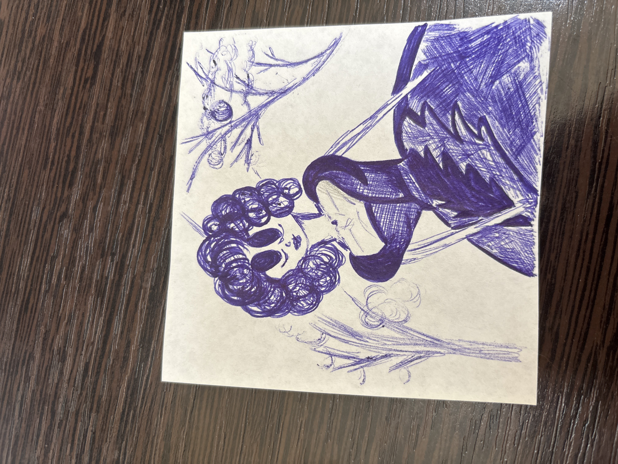 A drawing on a white, rectangular sheet of paper shows a stylized female figure with large, dark eyes and voluminous curly hair, wearing a dark, ornate dress. The figure is depicted in shades of purple and blue, with heavy hatching used to create texture and shading, particularly on the dress. The background includes abstract representations of trees and branches, also drawn in purple ink. The paper is placed on a dark, wooden surface with a prominent wood grain pattern. The drawing does not contain any recognizable landmarks or features that would indicate a specific location in Pervomayskiy, Russia.