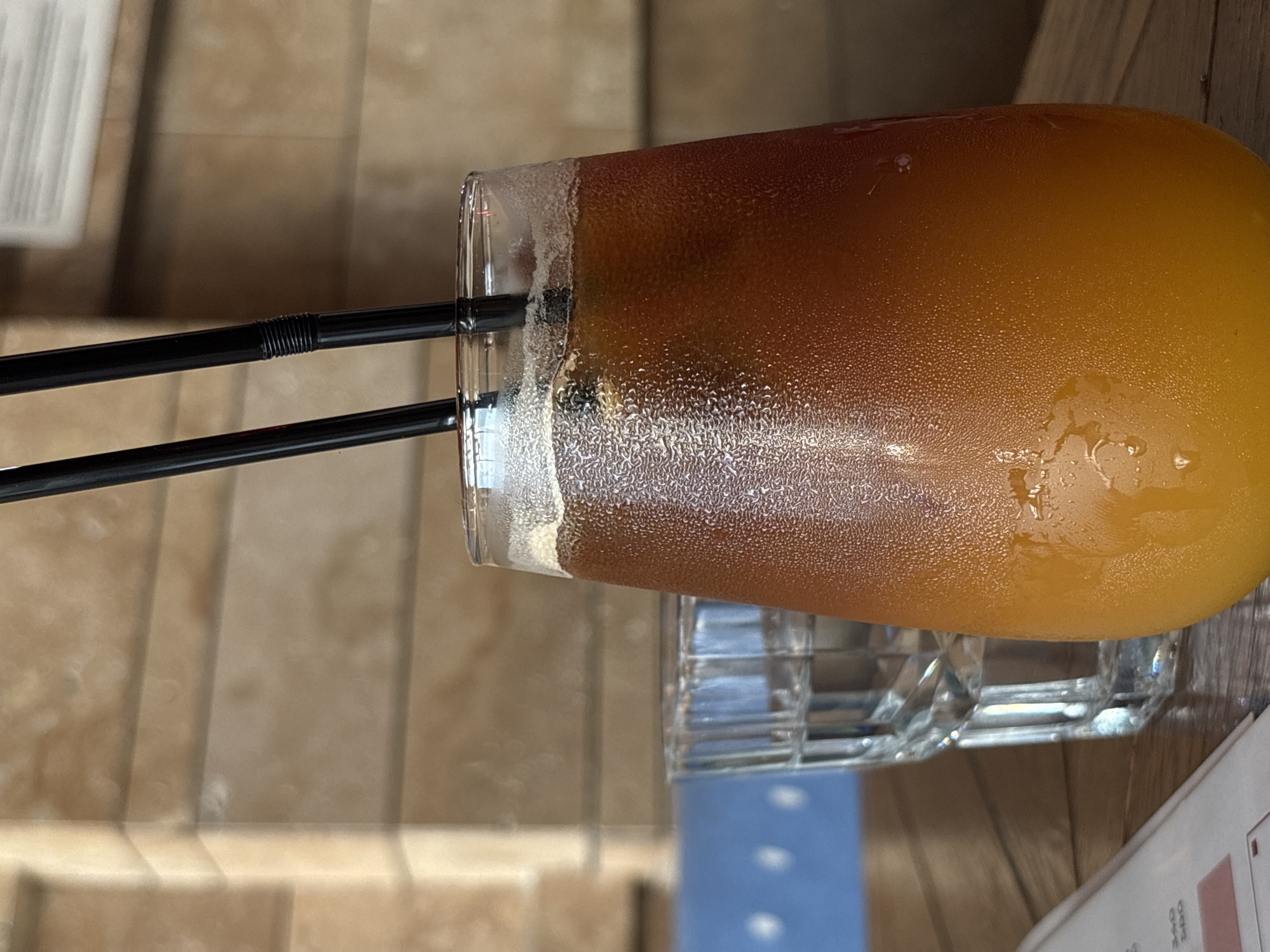An eye-level forensic view captured in the city of Perm, Russia, displays a close-up of two beverages on a wooden table. In the foreground, a tall, clear glass contains a layered, cold drink, dark brown at the top and orange-yellow at the bottom, with prominent condensation droplets coating its exterior. Two black plastic straws are inserted into the drink. Slightly behind and to the left of the main glass, a smaller, clear faceted glass holds water. The table surface is composed of light brown wooden planks. In the lower-left corner, a white menu or document with red accents is partially visible. The background features a light brown wall constructed of horizontal panels, with a section of a blue wall or object with white circular patterns visible to the left. A white rectangular sign is partially visible on the upper right background wall. No individuals are present in the frame.