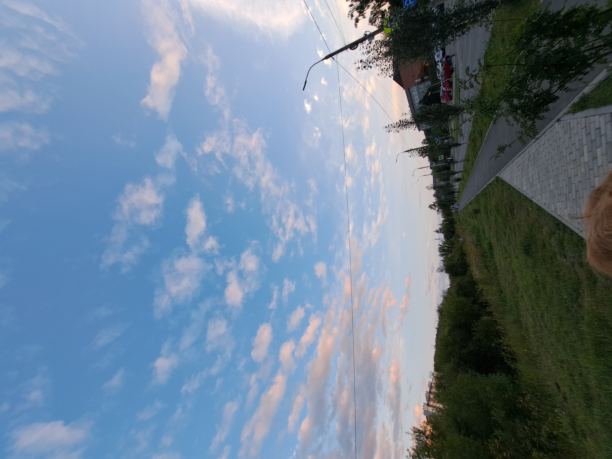 This image depicts a view of a street in Irkutsk, Russia, during what appears to be the late afternoon or early evening. The sky is a prominent feature, with a clear blue expanse dotted with scattered white and pinkish clouds, suggesting a soft, possibly sunset light.
In the foreground, a paved walkway made of grey rectangular stones runs diagonally across the frame. To the left of the walkway is a grassy slope, with overgrown green vegetation and some wild grass. Beyond the walkway, a two-lane asphalt road is visible, with several cars parked on the right side. The cars are predominantly red and white. There are also streetlights lining the road, and trees planted along its edge.
In the background, several buildings can be seen. On the right side, there are multi-story buildings with brick facades. On the left side, further in the distance, a modern residential building with a lighter facade is visible amidst lush green trees. A few people are present, walking along the sidewalk and the road. One individual is walking on the road, while others are on the walkway. The overall impression is a quiet urban scene, with elements of nature present alongside infrastructure. The lighting suggests a peaceful, possibly transitional time of day.
