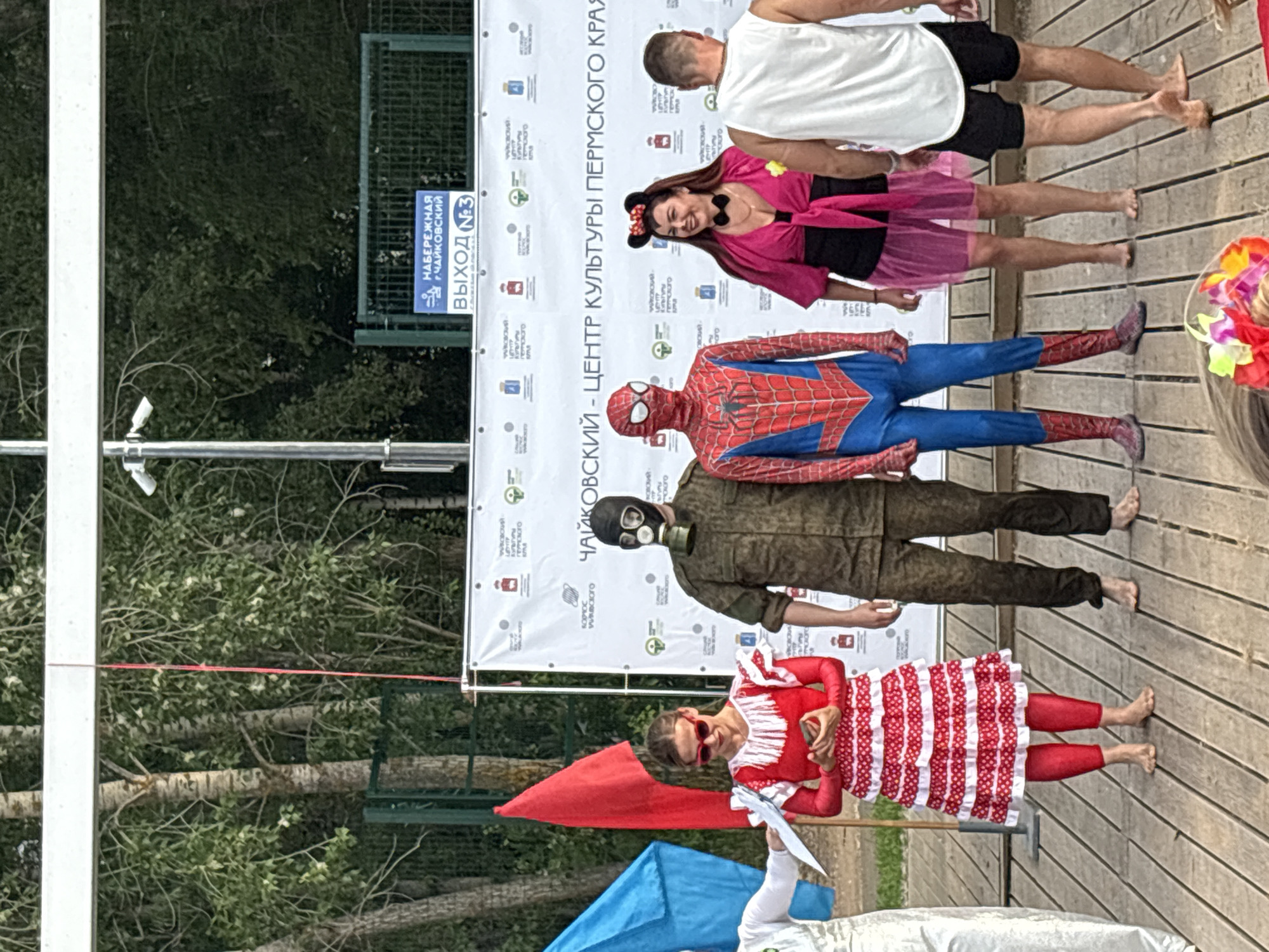 This image depicts an outdoor event in the city of Chaikovsky, Russia, likely at a cultural center. In the foreground, a group of people are standing on a wooden platform. On the left, a woman in a red and white polka dot dress and red tights is holding a mobile phone. To her right, a person in a full camouflage military uniform and a gas mask stands with their arms at their sides. Next to them, a person dressed as Spider-Man is posing, with their arms down. To the right of Spider-Man, a woman in a pink dress and Minnie Mouse ears is smiling and looking to her left. Further to the right, a man in a white tank top and black shorts is walking away from the camera, with his back to the viewer. Behind the group, a large white banner with text and logos is displayed. Above the banner, a sign in blue reads "НАБЕРЕЖНАЯ г. ЧАЙКОВСКИЙ" which translates to "Embankment of Chaikovsky City". Below that, another sign indicates "ВЫХОД №3" meaning "Exit No. 3". Trees and foliage are visible in the background, along with a green fence and security cameras mounted on a pole. Two flags, one red and one blue, are also visible on the left side of the image.