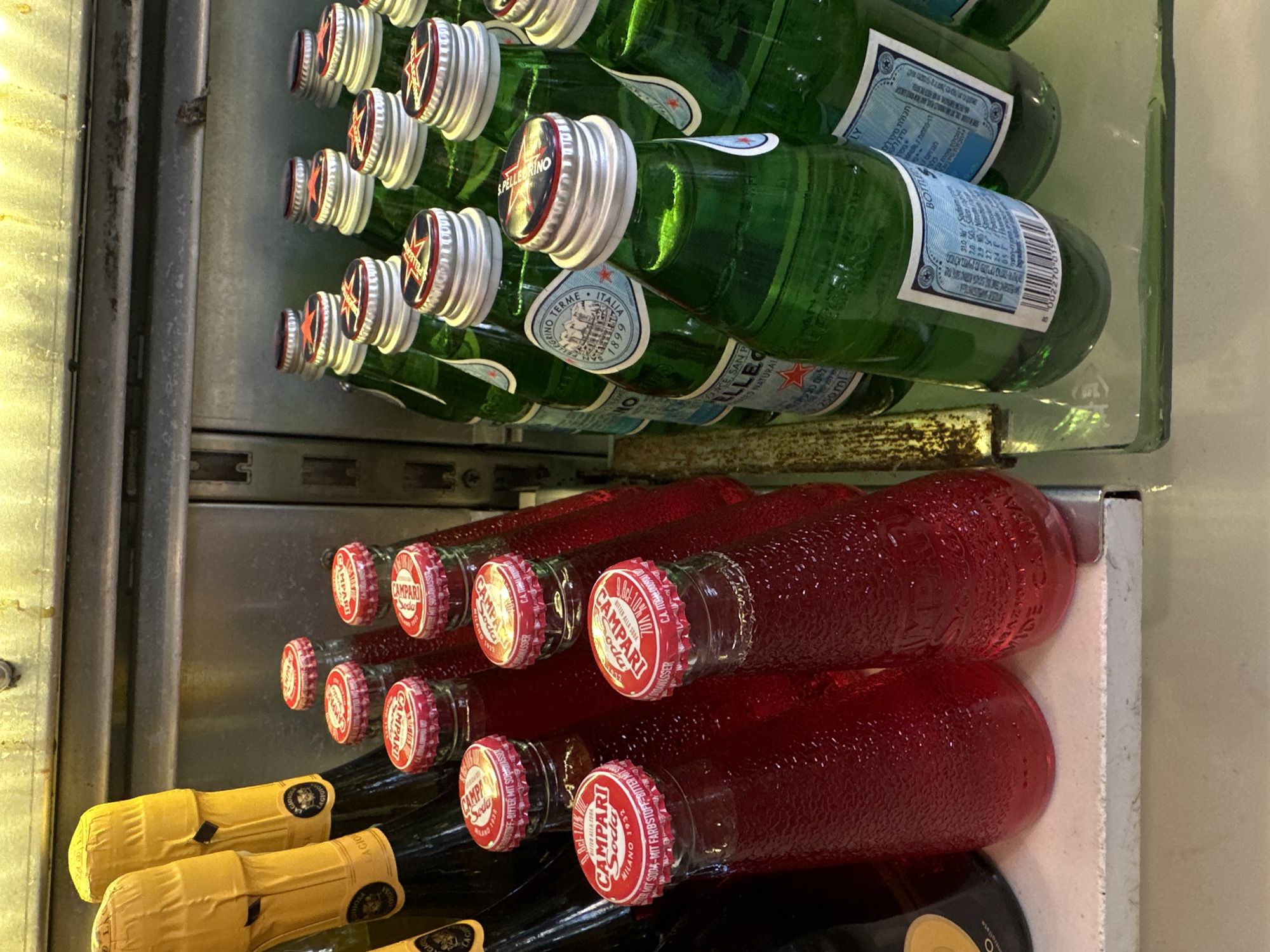 The image shows a close-up, vertical shot of bottled beverages displayed in a refrigerator or on shelves. The top section features several green glass bottles with silver caps, primarily San Pellegrino sparkling water, evidenced by the visible labels with "PELLEGRINO," "TERME," and "ITALIA." The bottles are stacked, and some labels include text like "250ml" and "BOTTLE."
The lower section of the image displays rows of red liquid in clear glass bottles with red caps. These are identifiable as Campari Soda bottles, with the "CAMPARI Soda" logo clearly visible on the caps, along with text indicating "MILANO 1932" and "BITTER MIT SODA-WASSER." There are also two bottles with yellow foil wrappers on their necks, likely wine bottles. The setting appears to be a commercial establishment, possibly a bar or store, given the organized display of beverages. The lighting suggests an indoor environment, potentially during the day or under artificial light. There are no people visible in the image. The overall impression is a curated selection of drinks ready for consumption.