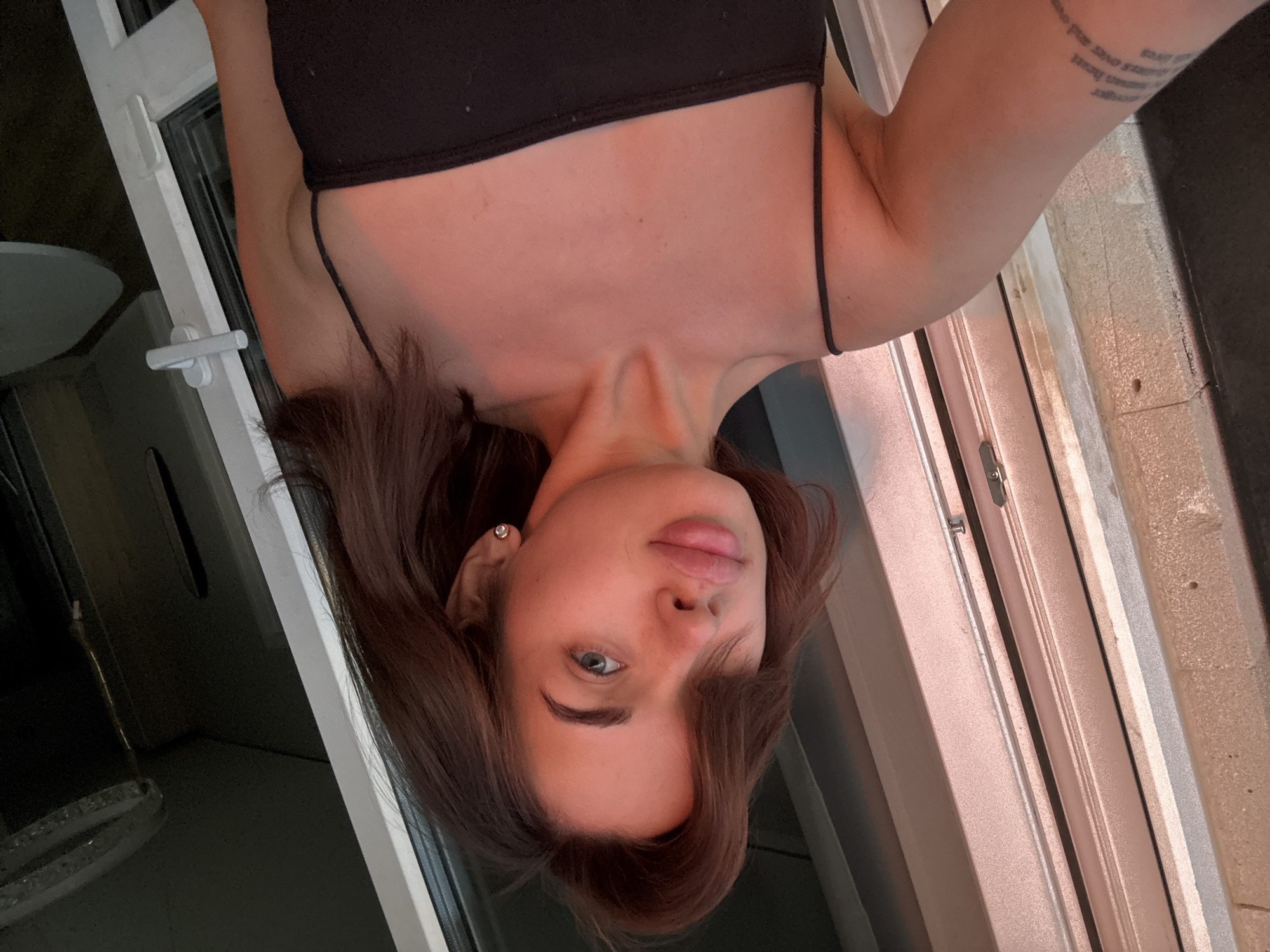 A woman wearing a black tank top is positioned near a window, seemingly taking a selfie. She has dark hair and light eyes. The scene appears to be indoors, with a view of the city of Лобаново, Russia visible outside the window. The precise location within the city is unclear.