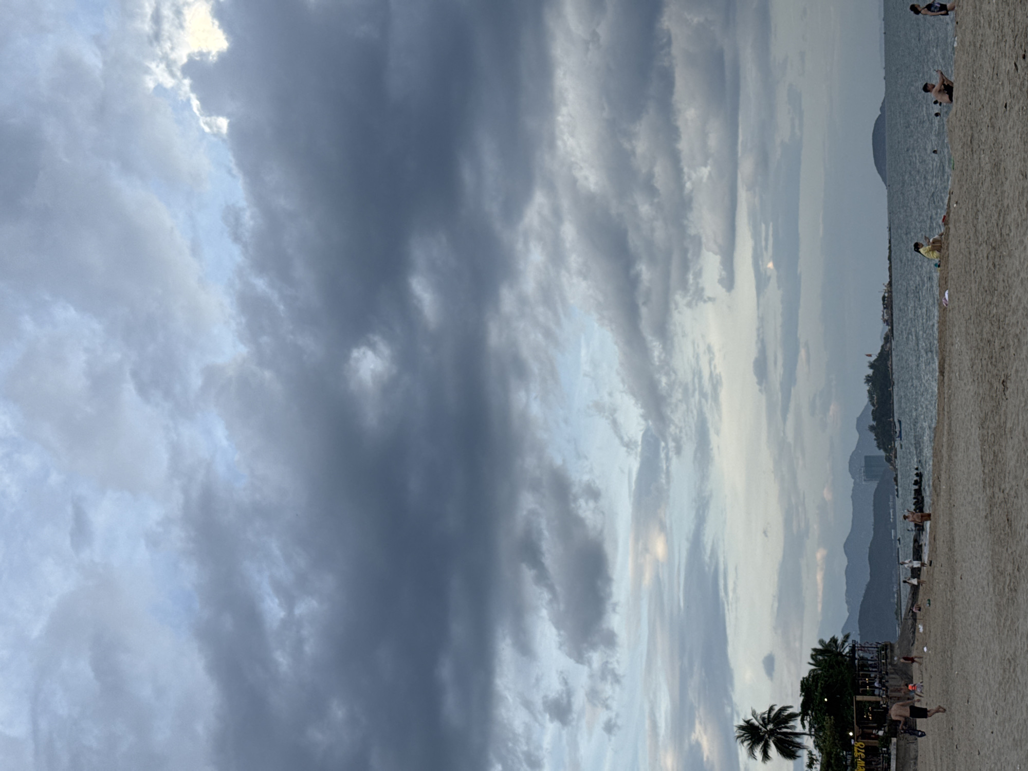 The image depicts a beach scene in Nha Trang, Vietnam, under a cloudy sky. The foreground is a sandy beach with several people scattered across it. On the left side of the beach, there is a palm tree and a building with signage that reads "View 378." The middle ground shows the sea with a few small boats. In the background, there are mountains and islands. The overall mood of the image is serene, with the overcast sky contributing to a slightly muted atmosphere.